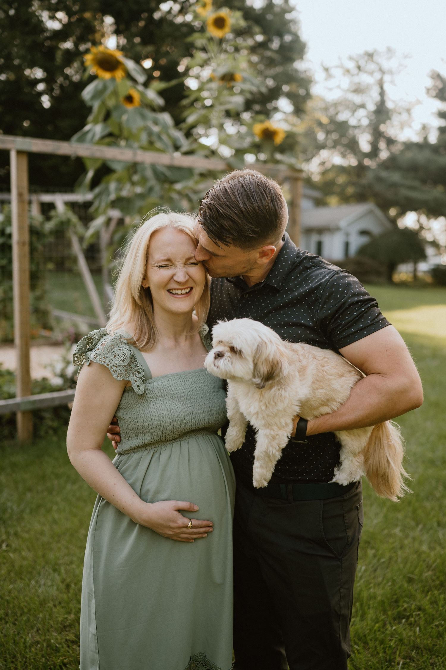 A couple poses with their small white dog in a backyard garden with sunflowers.