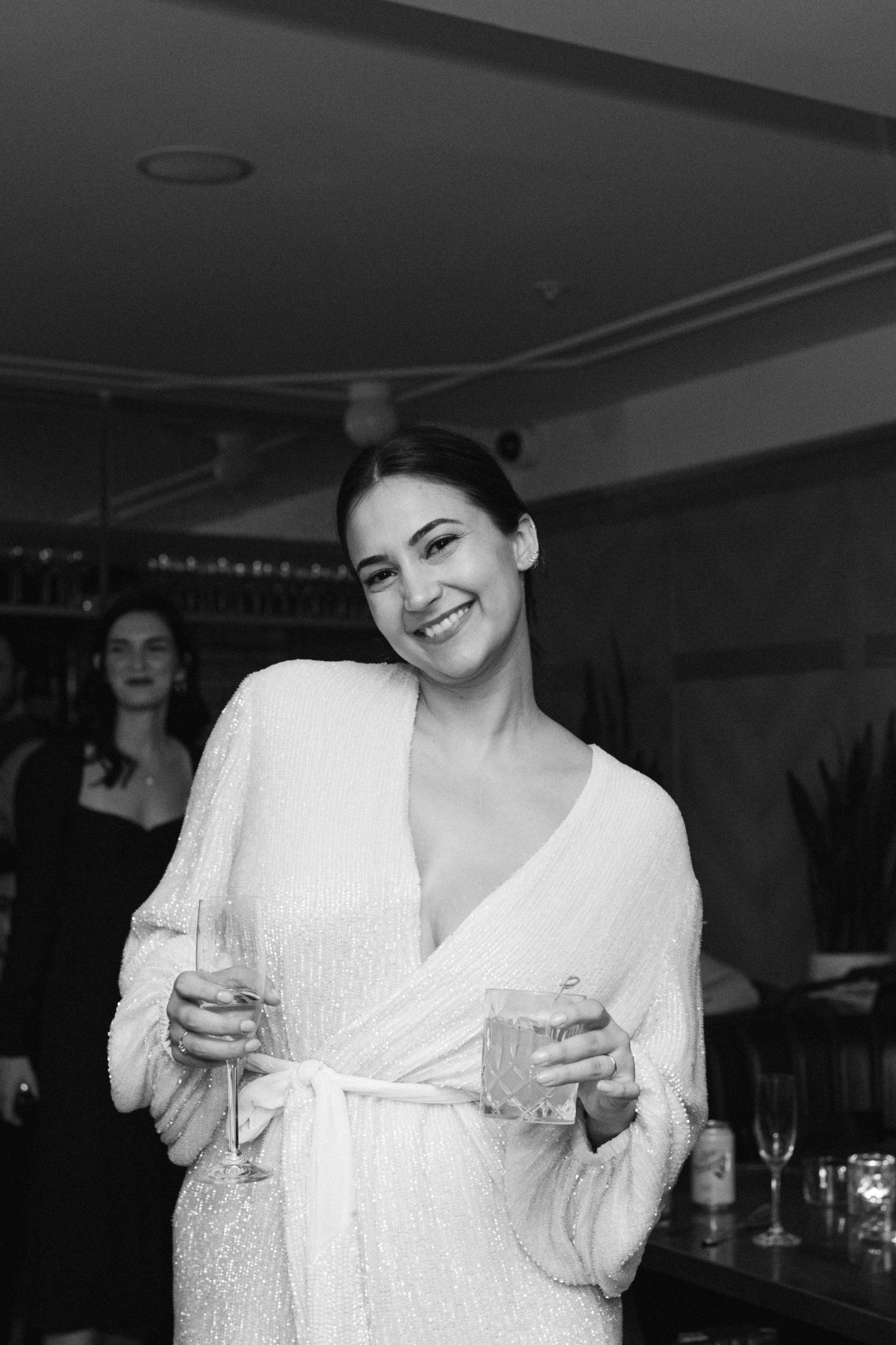 A woman in white holding champagne glasses smiles during a celebration.