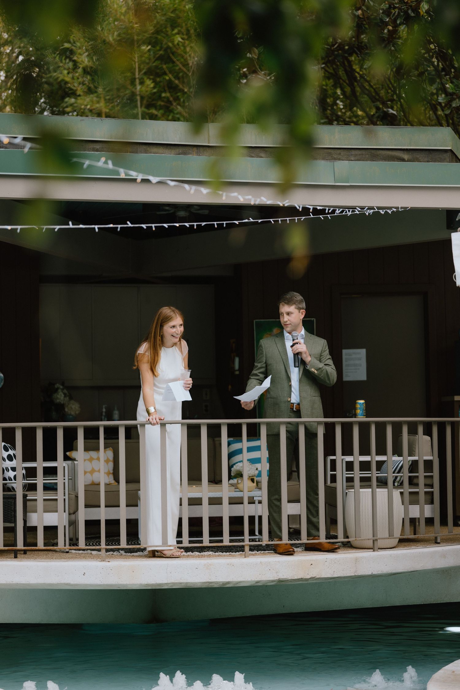 Two people standing on a covered deck overlooking a pool during a formal event.