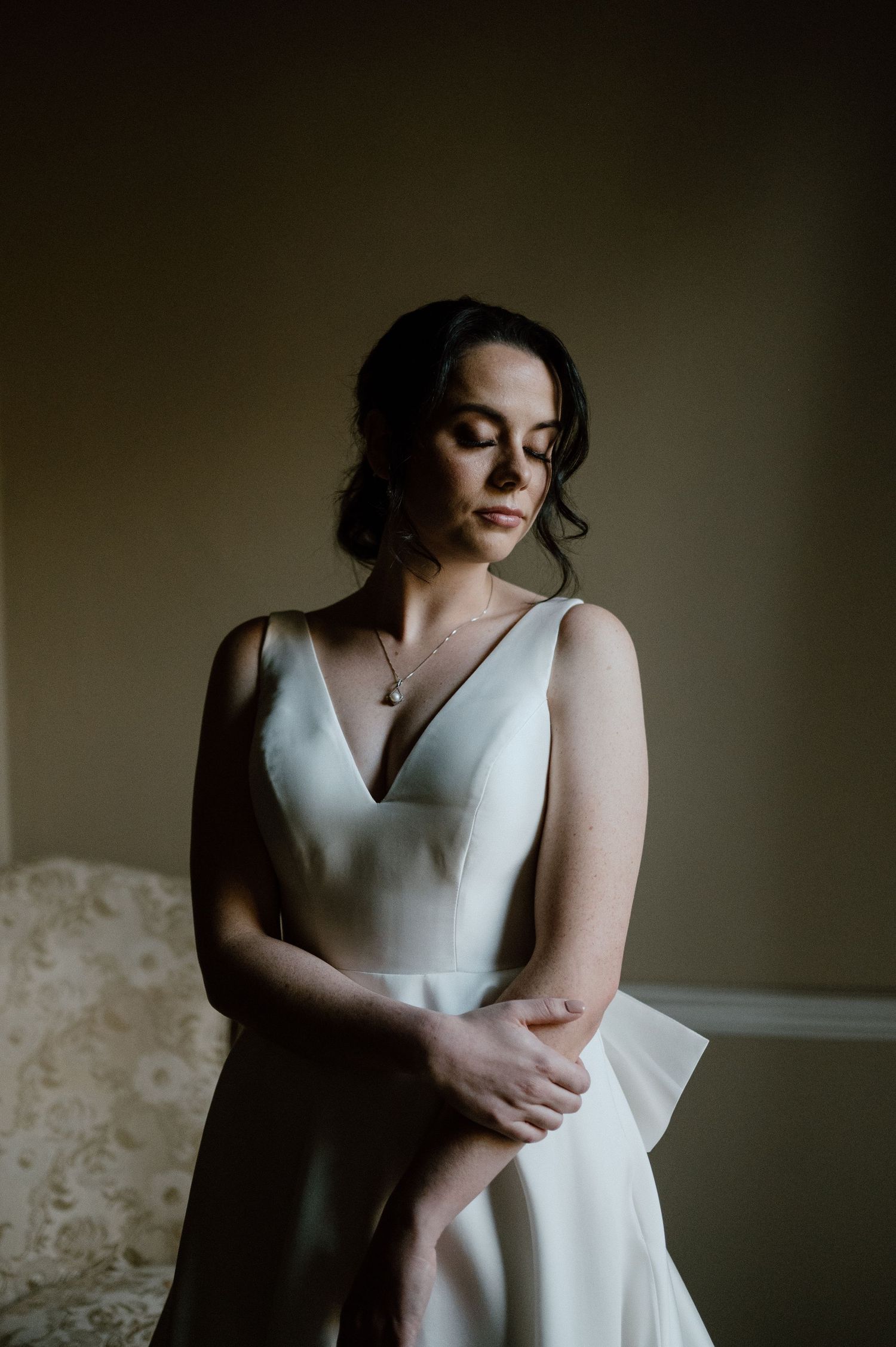 Moody portrait of someone in a sleek white wedding dress with V-neckline in soft window light.