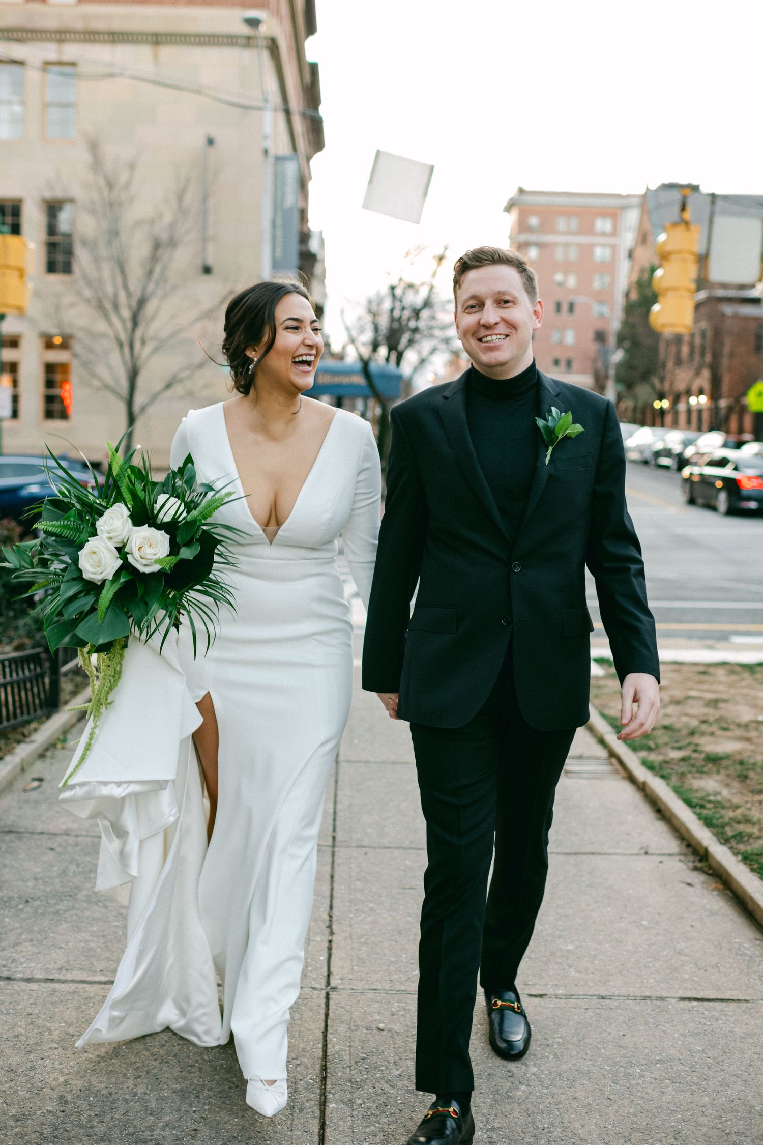 A joyful couple walks down a city sidewalk on their wedding day, with the bride carrying white flowers.