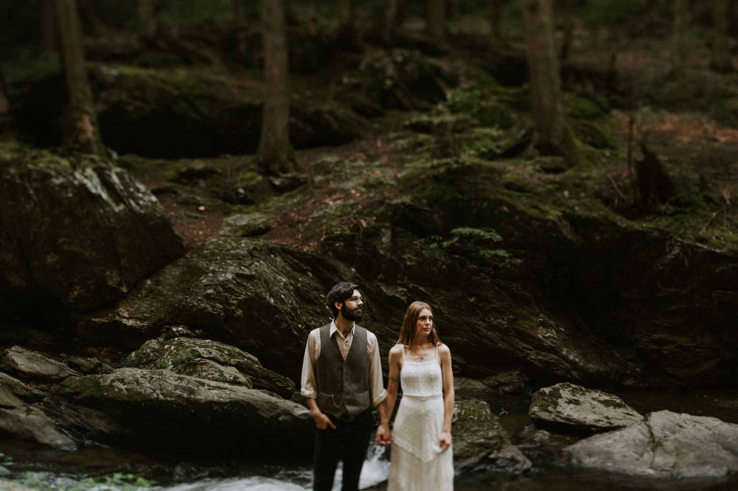 A couple explores a rocky forest landscape near a stream.