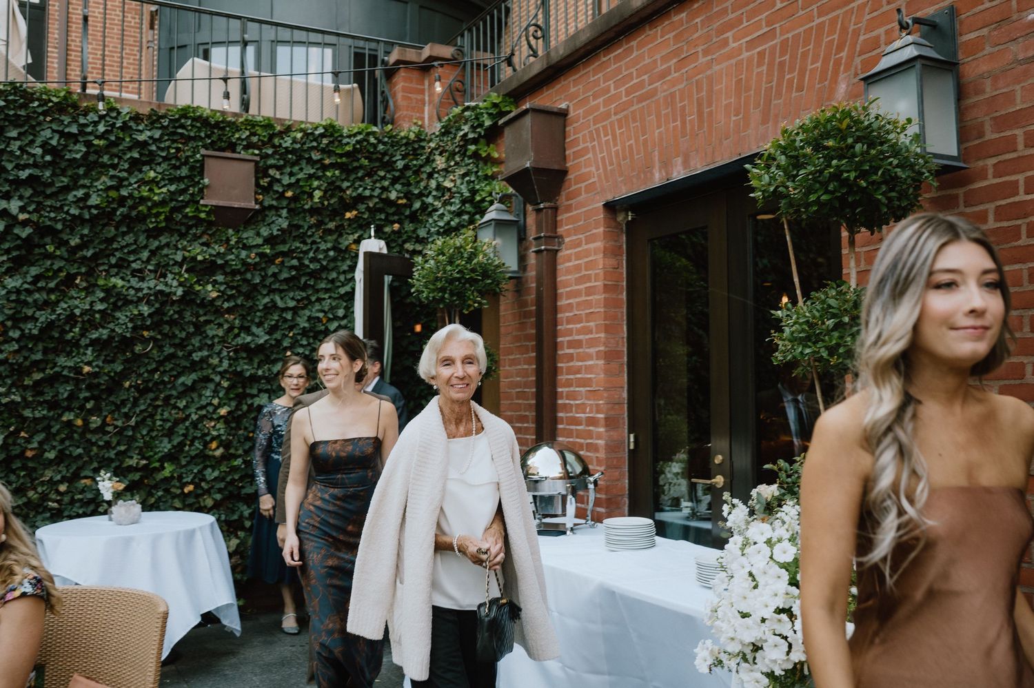 Guests mingling at an outdoor reception with ivy-covered brick walls and white tables.