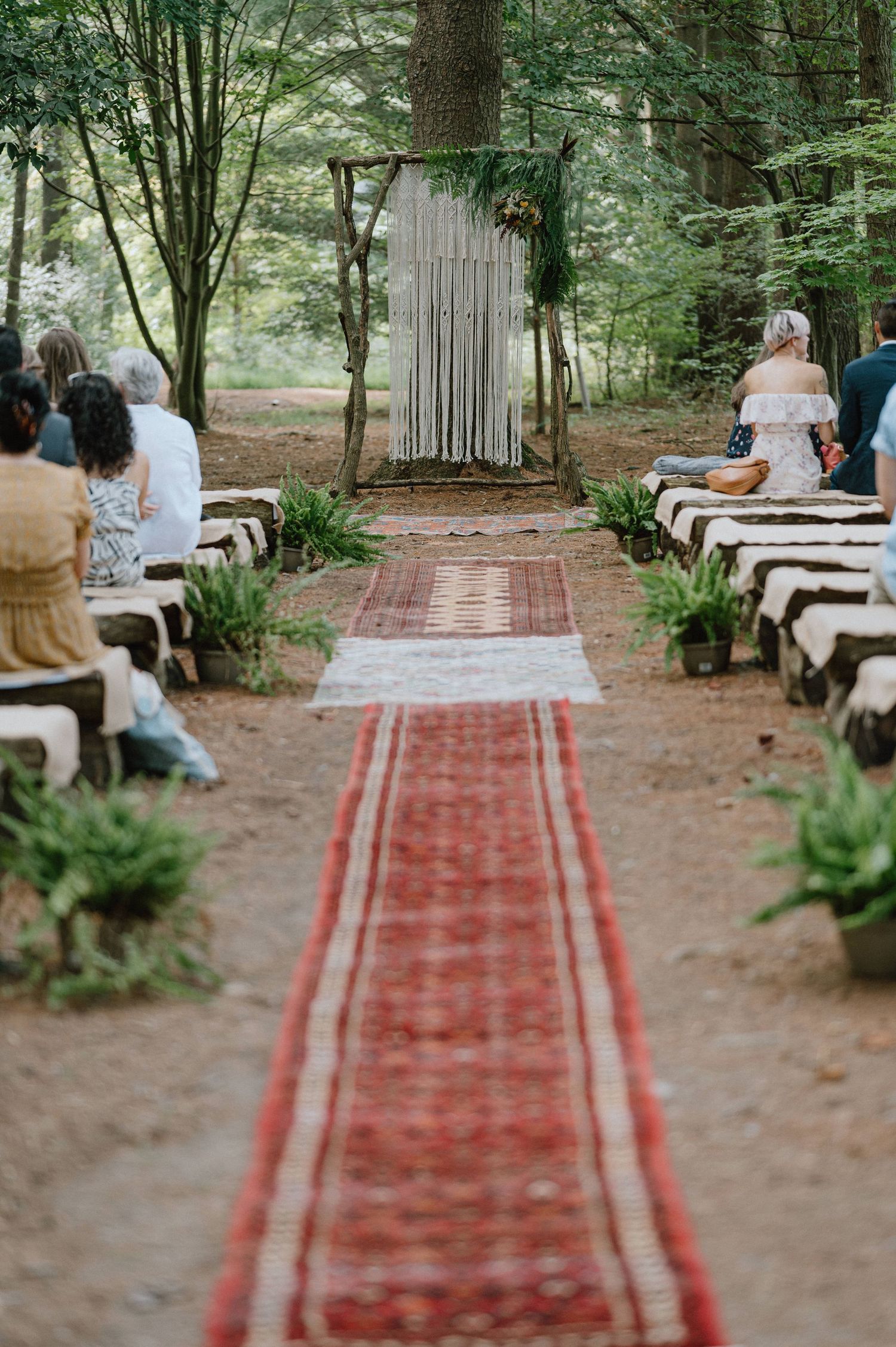 An outdoor forest wedding ceremony setup with a red carpet aisle and rustic white fabric backdrop.
