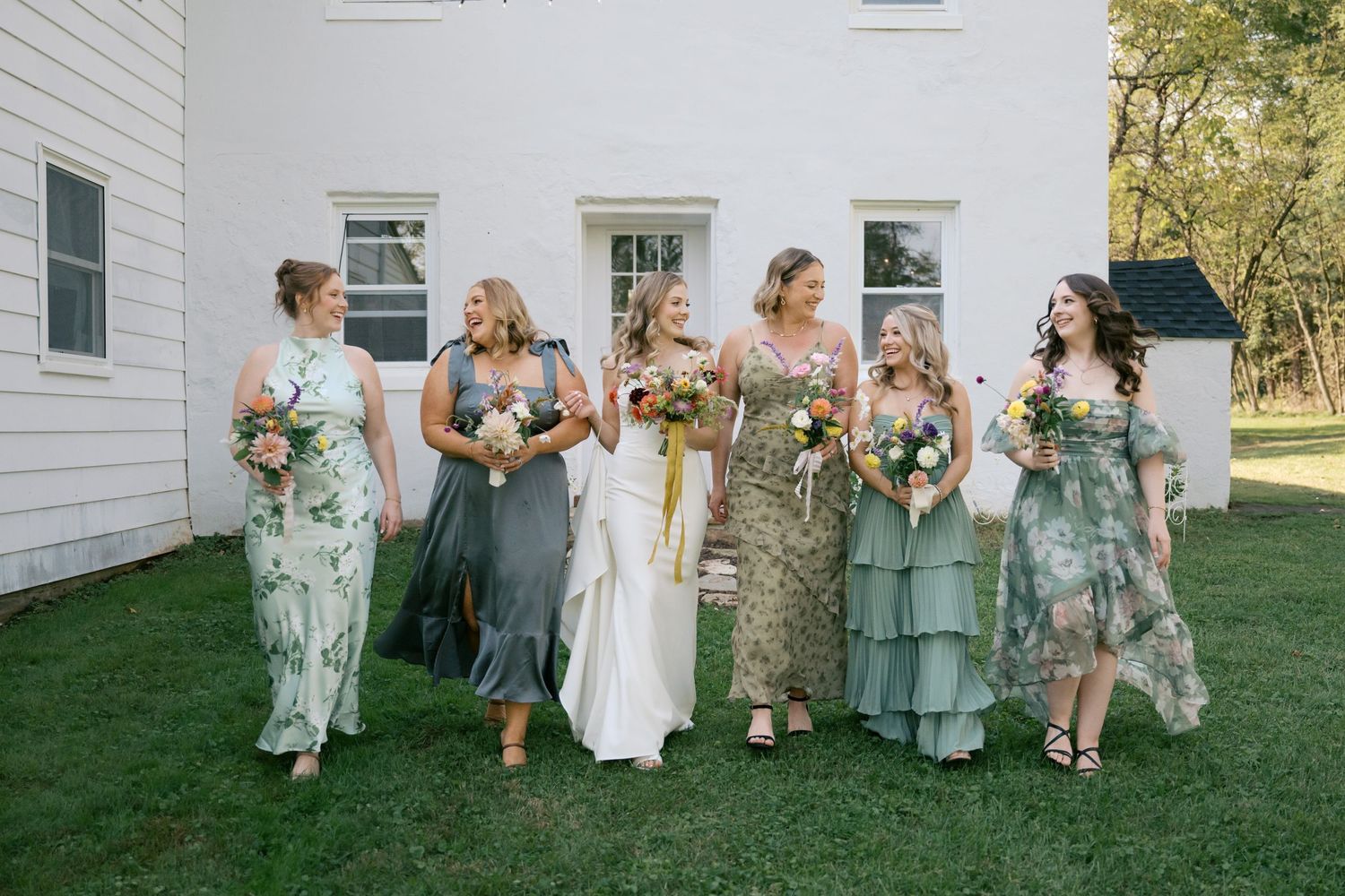 Wedding party in green floral dresses laughing together against white building backdrop.