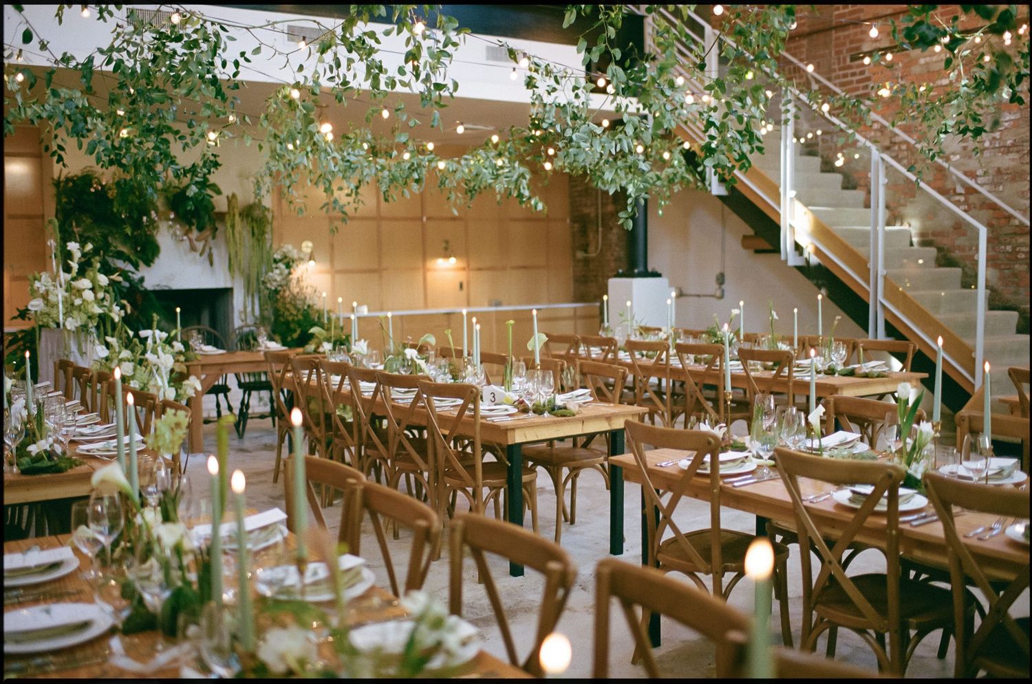 Wedding reception tables arranged under string lights with natural greenery and wooden chairs.