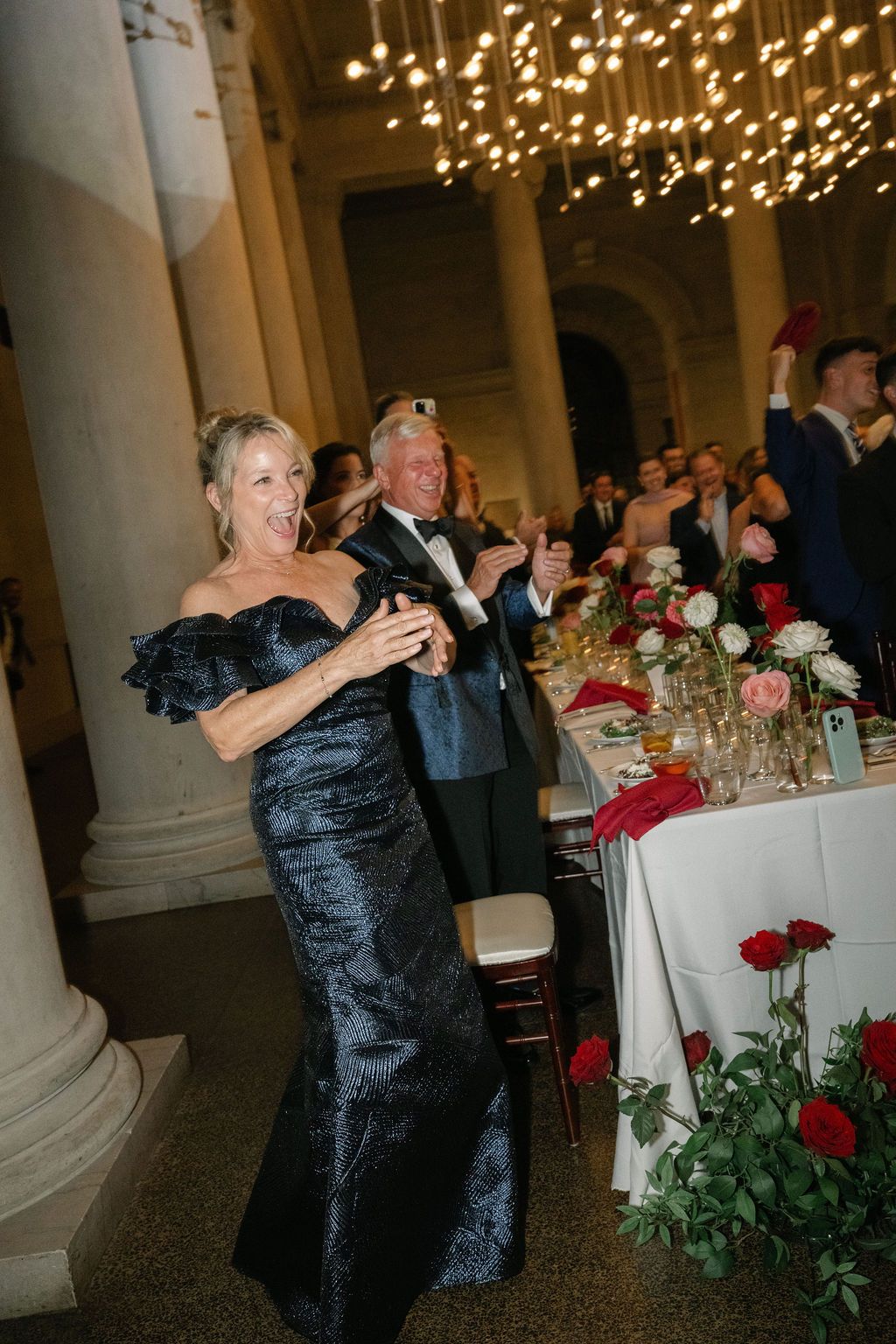 A couple dances together at an elegant wedding reception with string lights and red rose centerpieces decorating the venue.