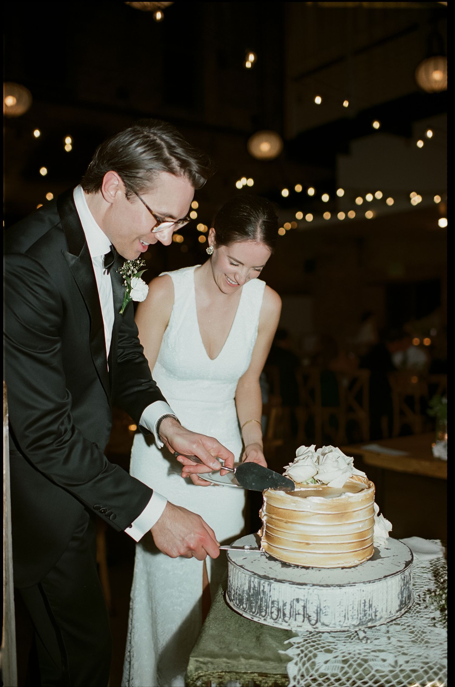 A couple cuts into their elegant gold-rimmed wedding cake together under twinkling reception lights.