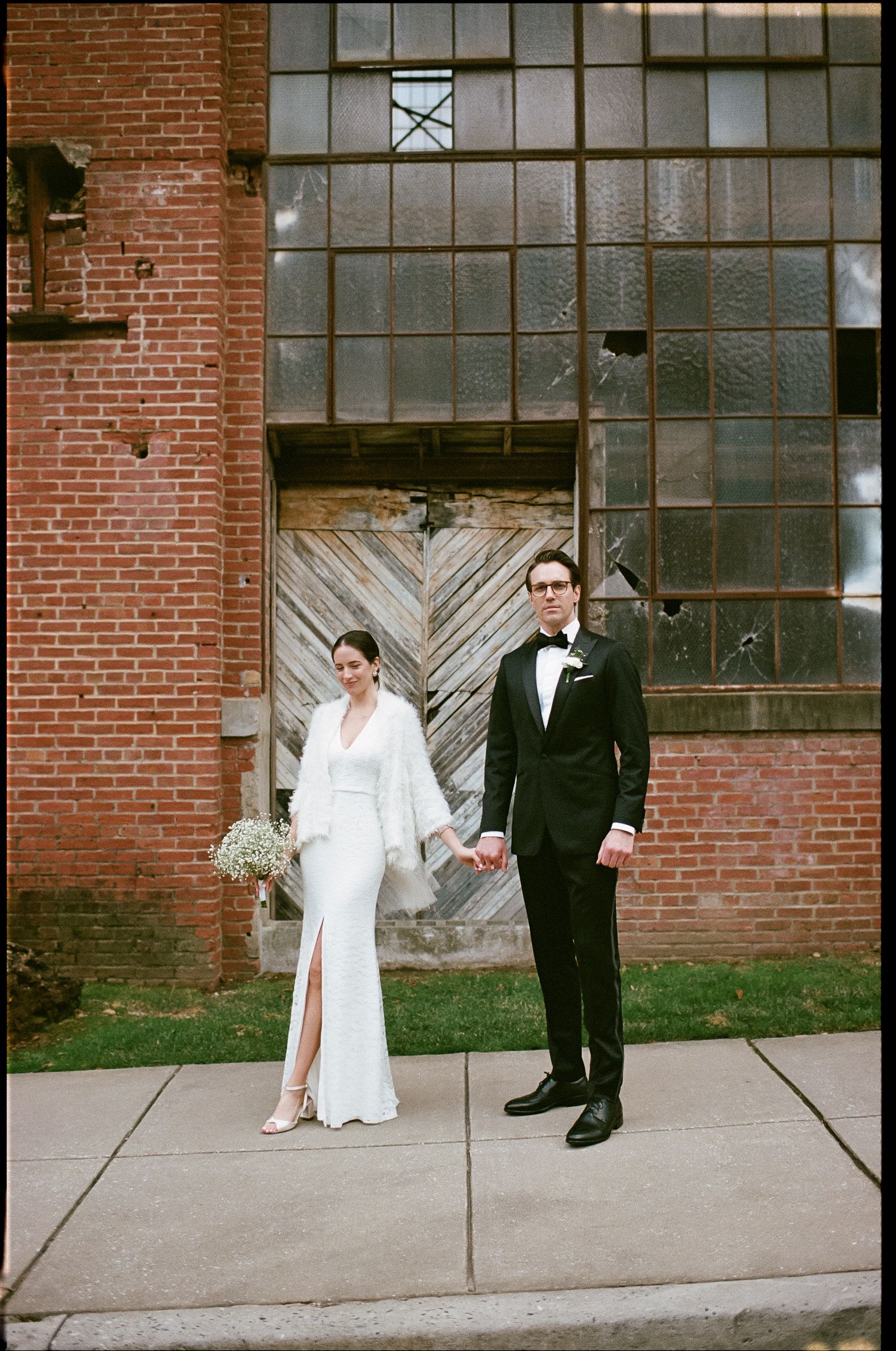 A stylish couple poses against an industrial brick building with large vintage windows.