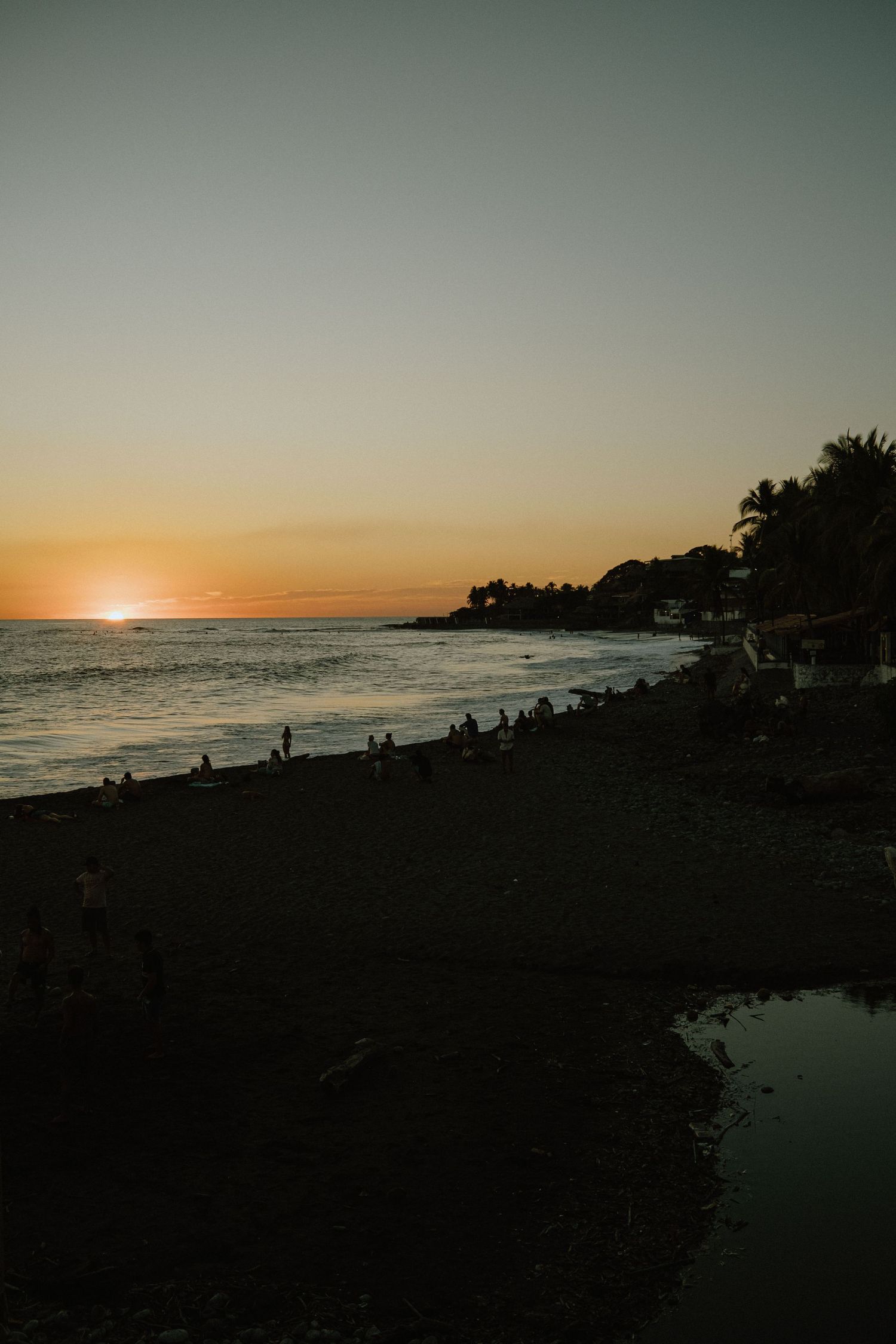 Dramatic orange sunset over a beach with silhouettes of people and palm trees against the darkening sky.