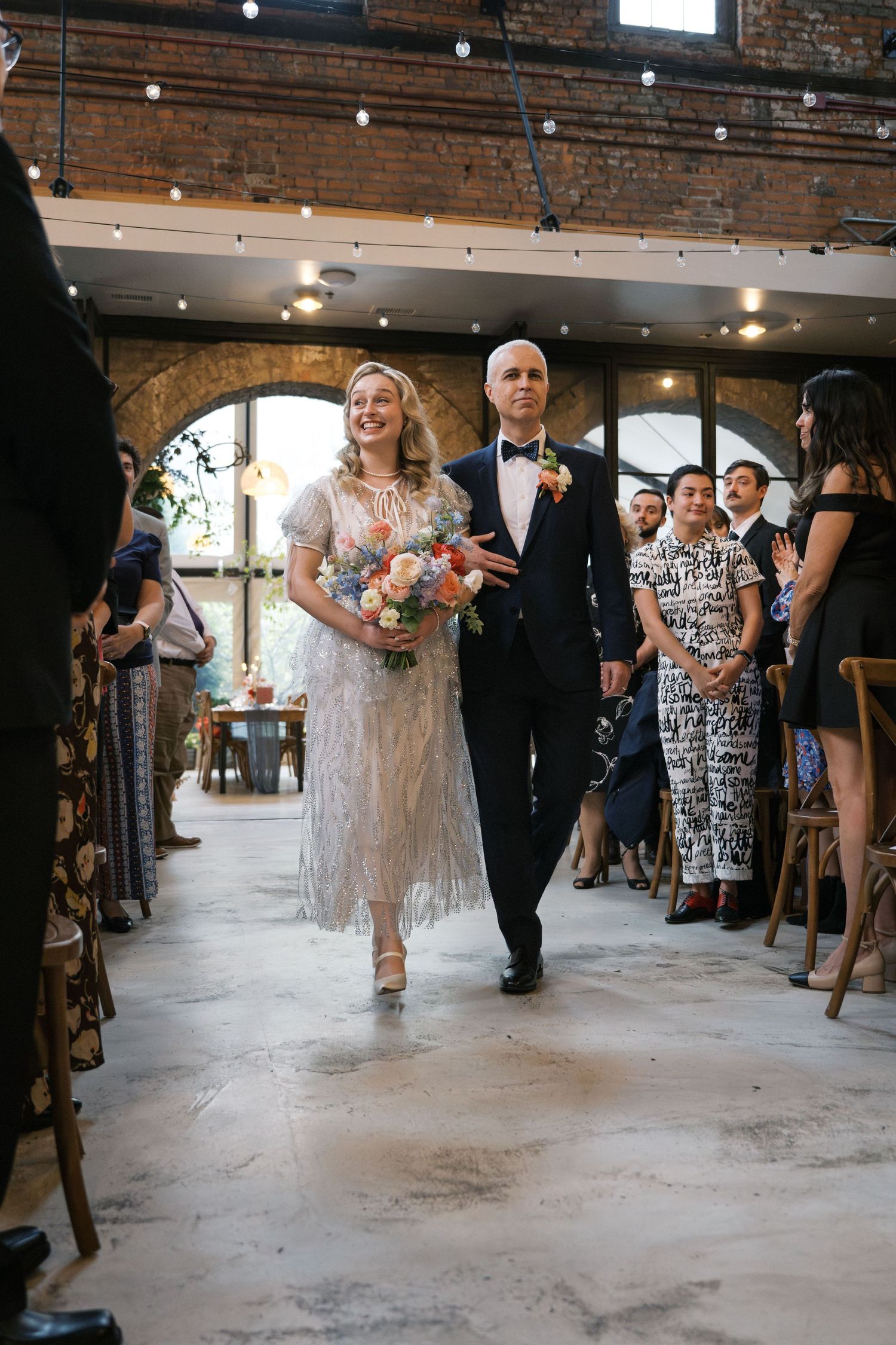 Couple walks down the aisle at rustic industrial wedding venue with exposed brick walls and string lights overhead.