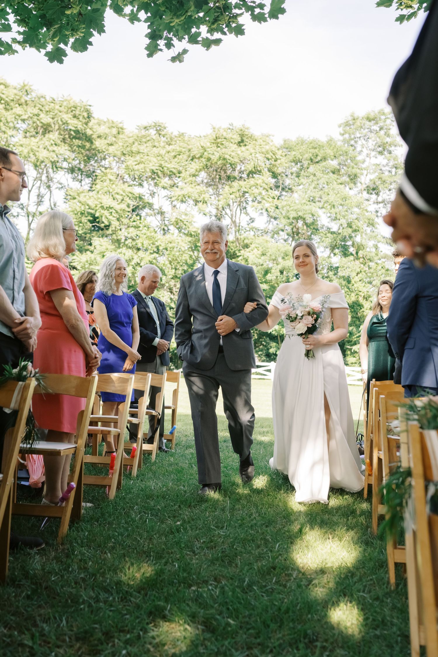 Wedding ceremony processional on lawn with wooden chairs and guests in summer attire.