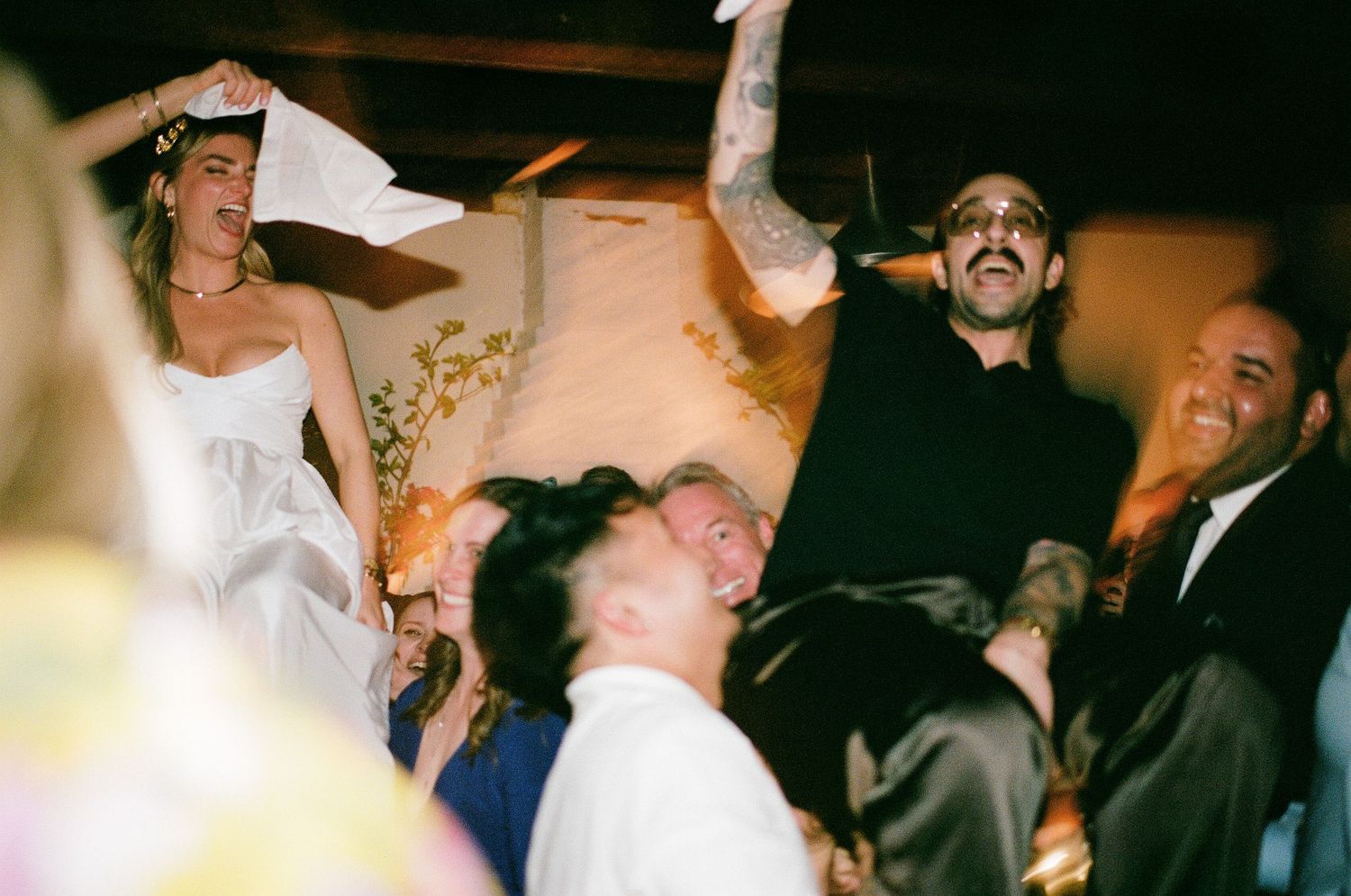 Guests celebrating joyfully at a wedding reception with people being lifted in the air during lively dance festivities.