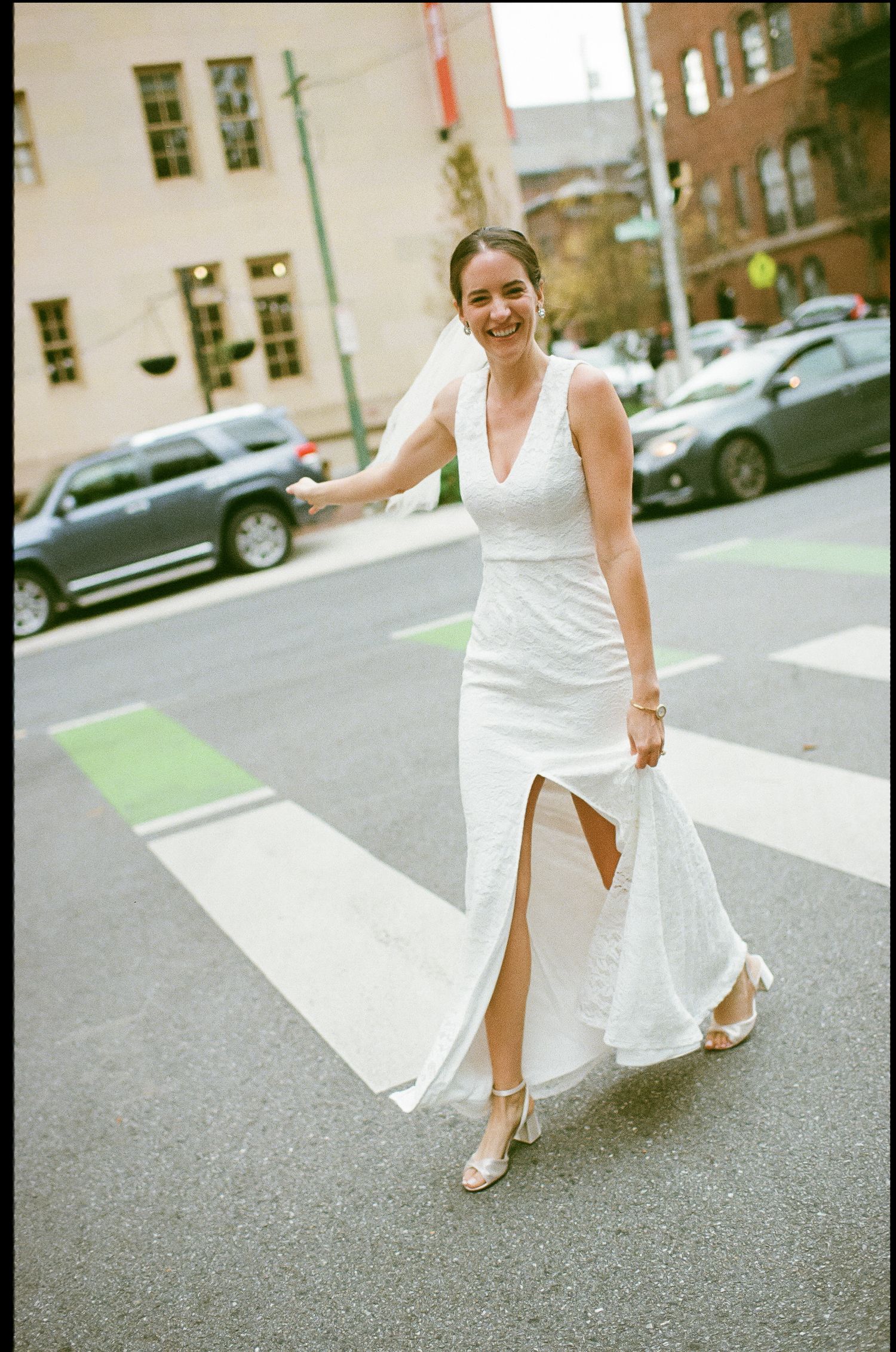 Bride in sleek white dress with slit walks across crosswalk on city street.