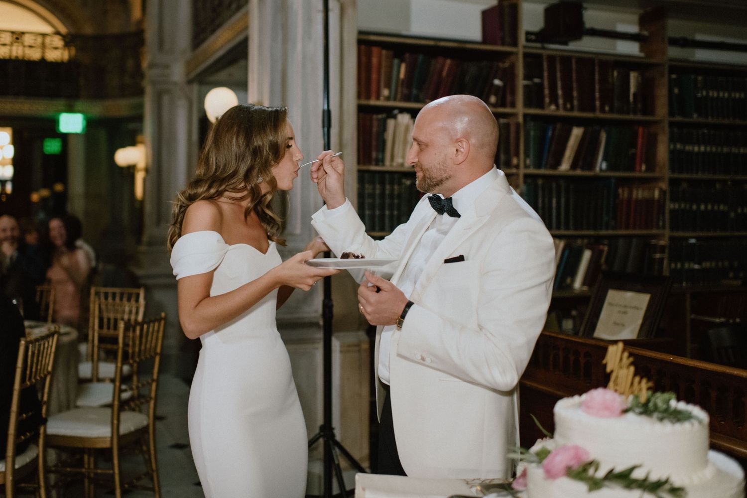 Two people in white formal attire share an intimate moment while cutting their wedding cake in an elegant library setting.