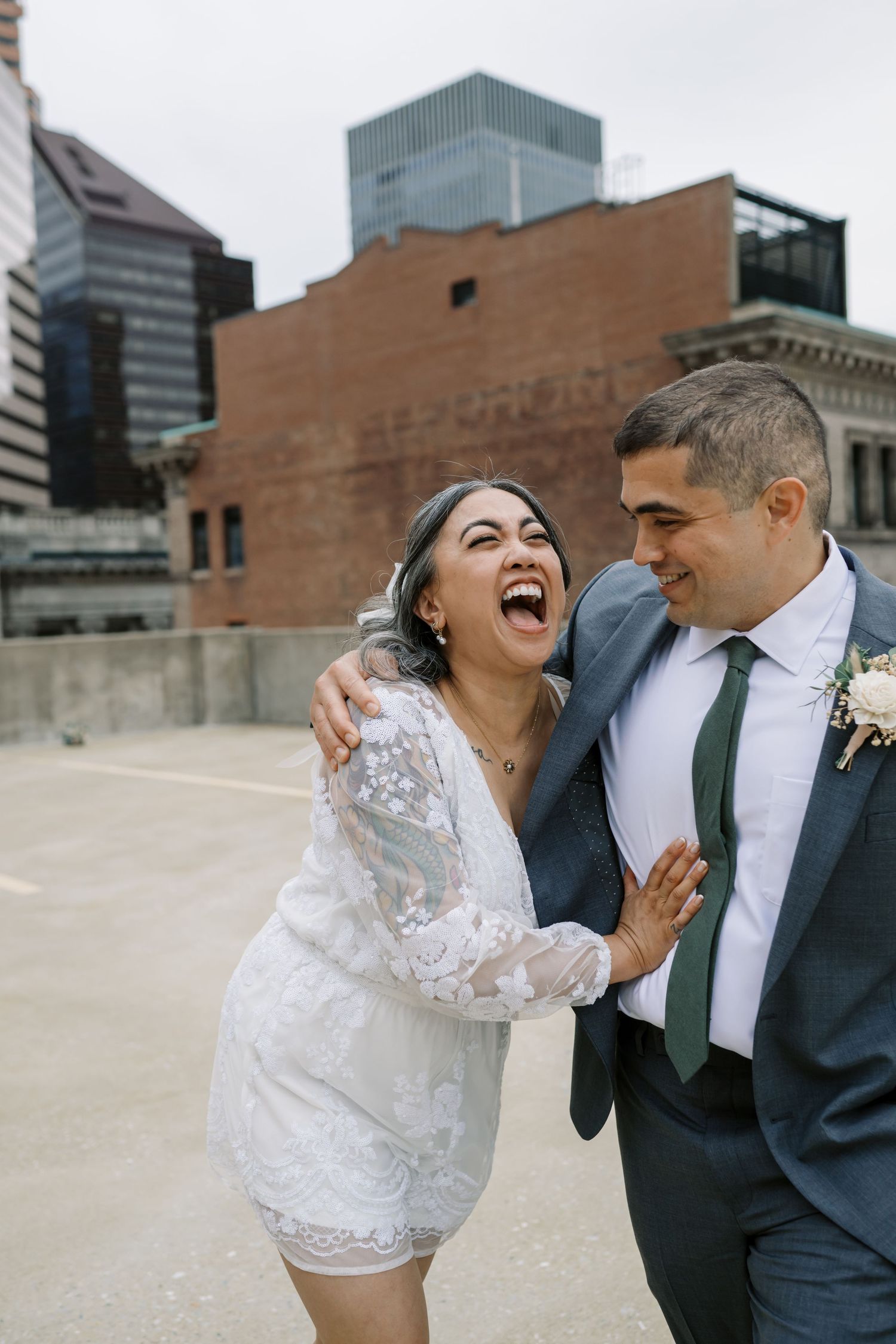 A couple shares a candid laughing moment against an urban backdrop with buildings.