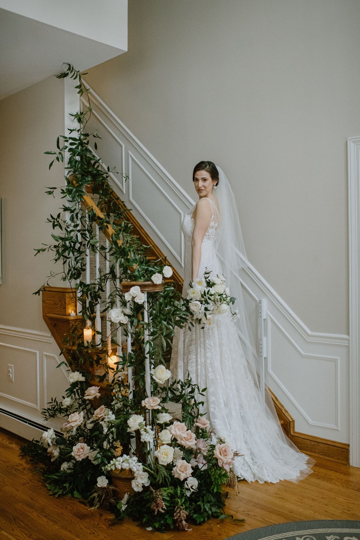 A bride poses on a staircase adorned with lush floral arrangements and candlelight.