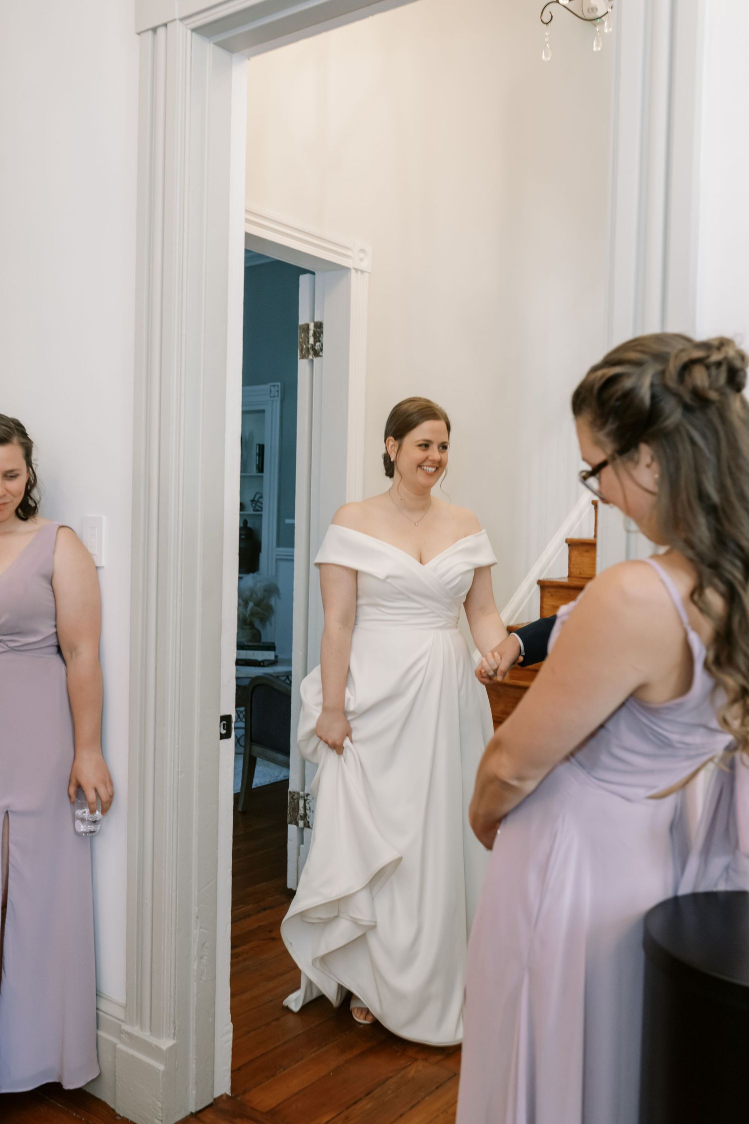 Wedding party members in lavender dresses gather in a bright indoor hallway.
