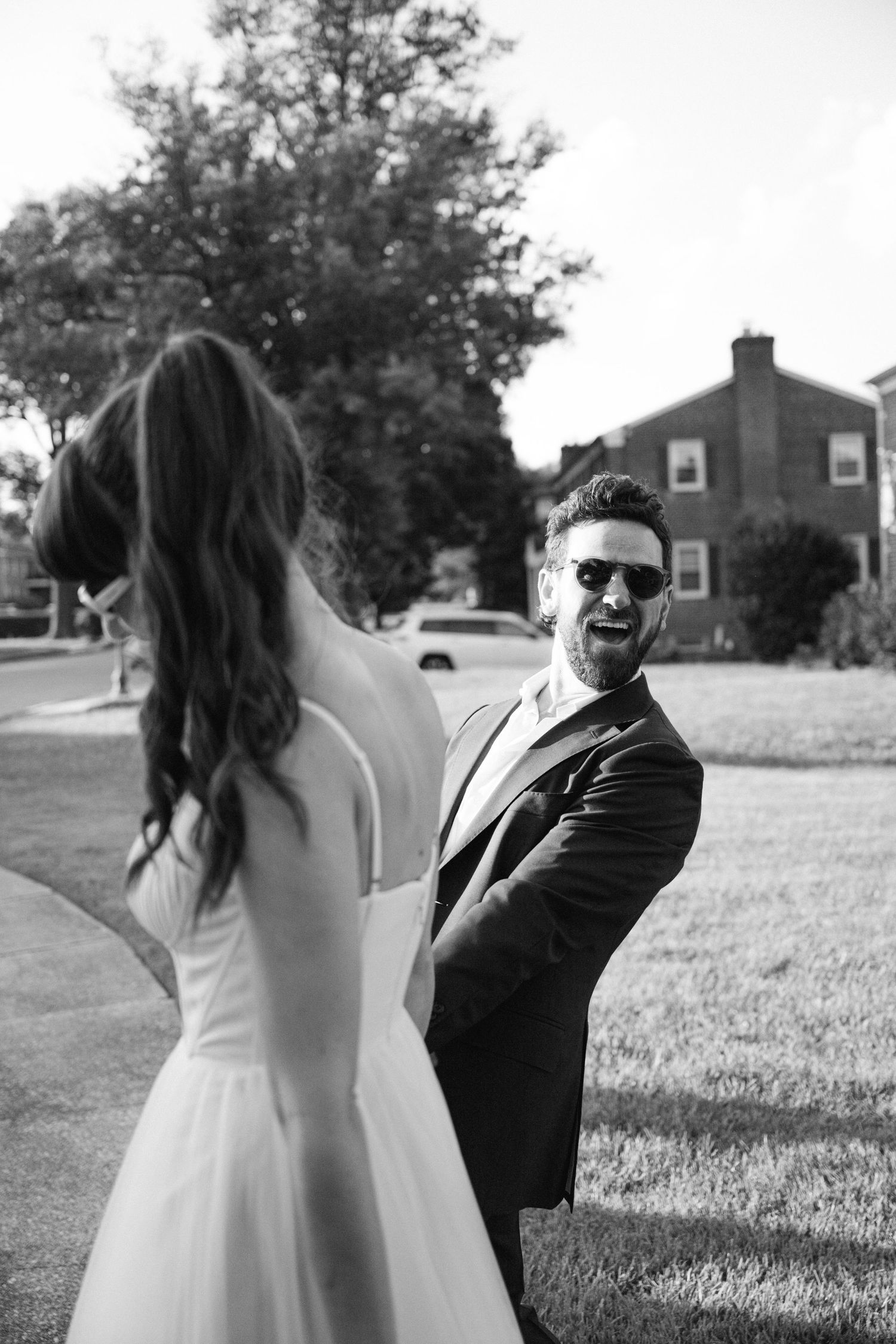 A black and white wedding photo shows a couple sharing a joyful moment outdoors with suburban homes in background.
