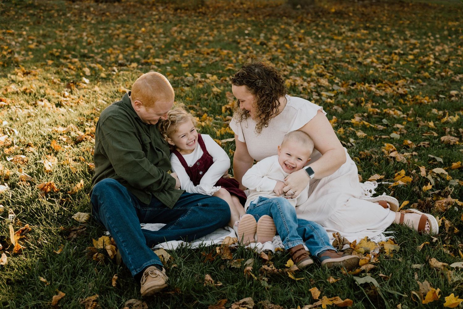 Family relaxing together on fallen autumn leaves during sunset photoshoot.