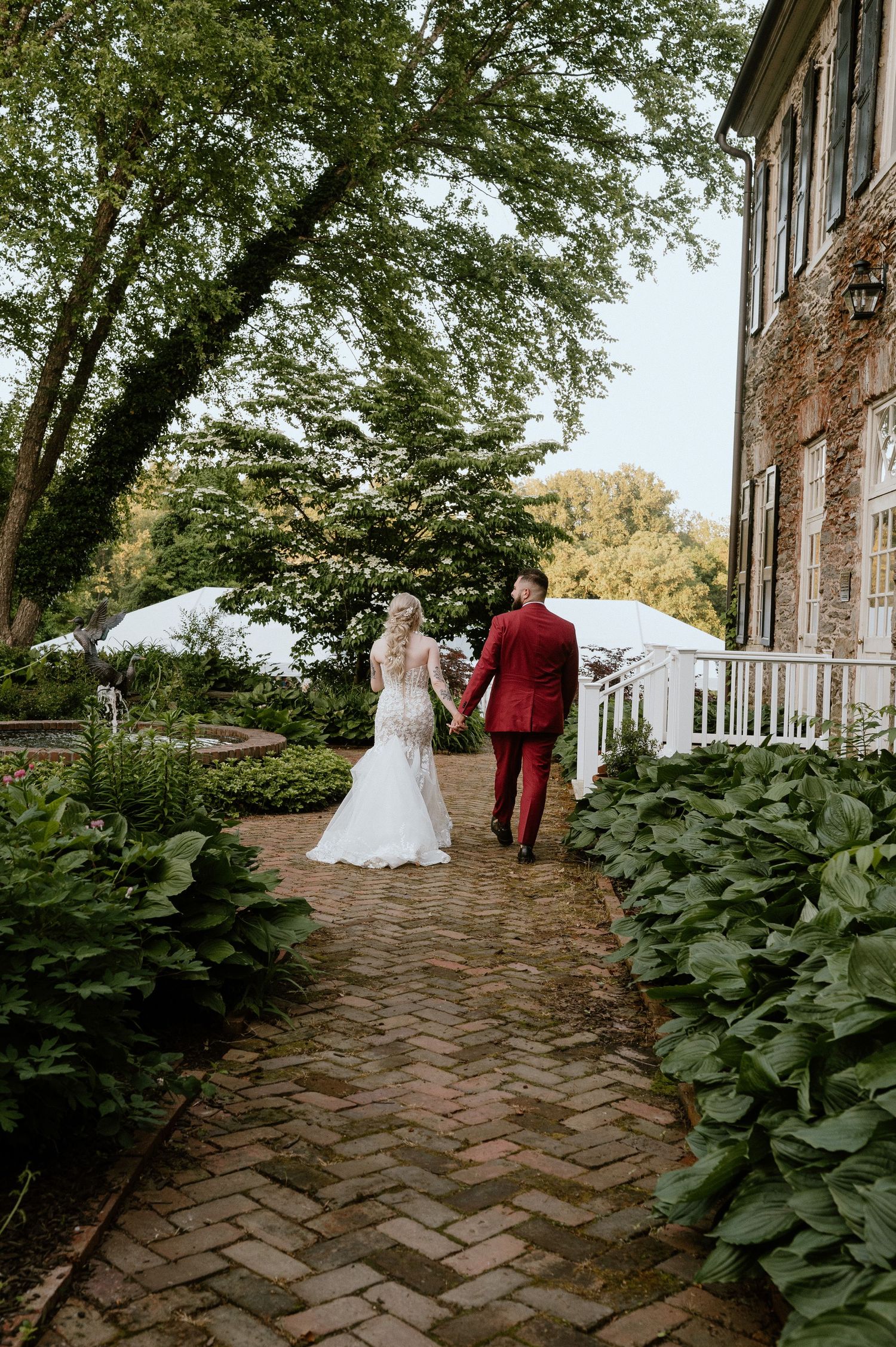 A couple walks along a brick pathway beside lush greenery and a historic stone building.