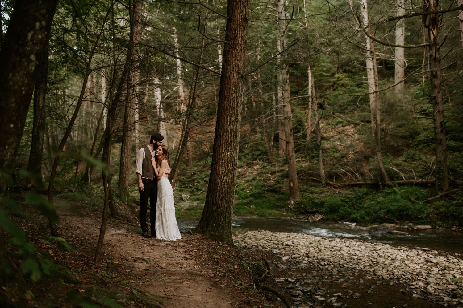 A couple stands together on a forest path beside a peaceful stream in moody woodland lighting.