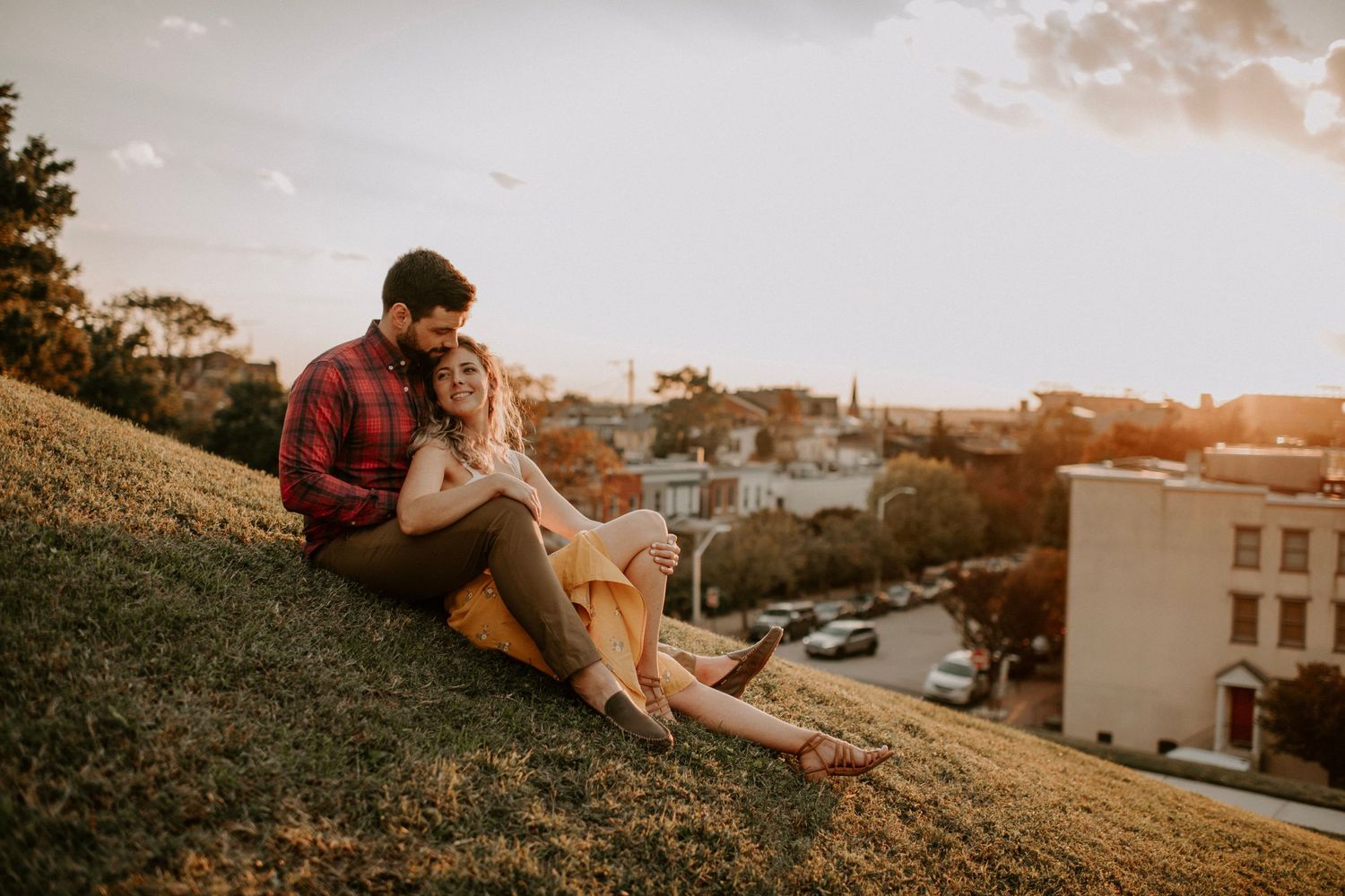Couple sits together on grassy hill overlooking urban landscape at sunset.