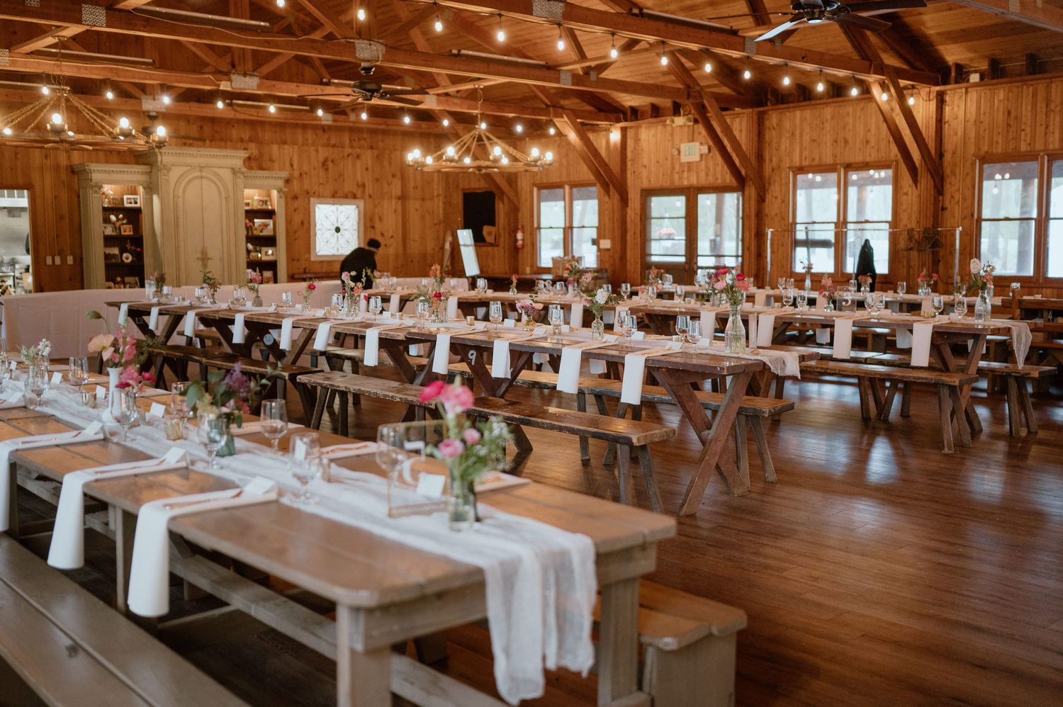 A rustic wedding reception venue with long wooden tables, white linens, and string lights overhead.