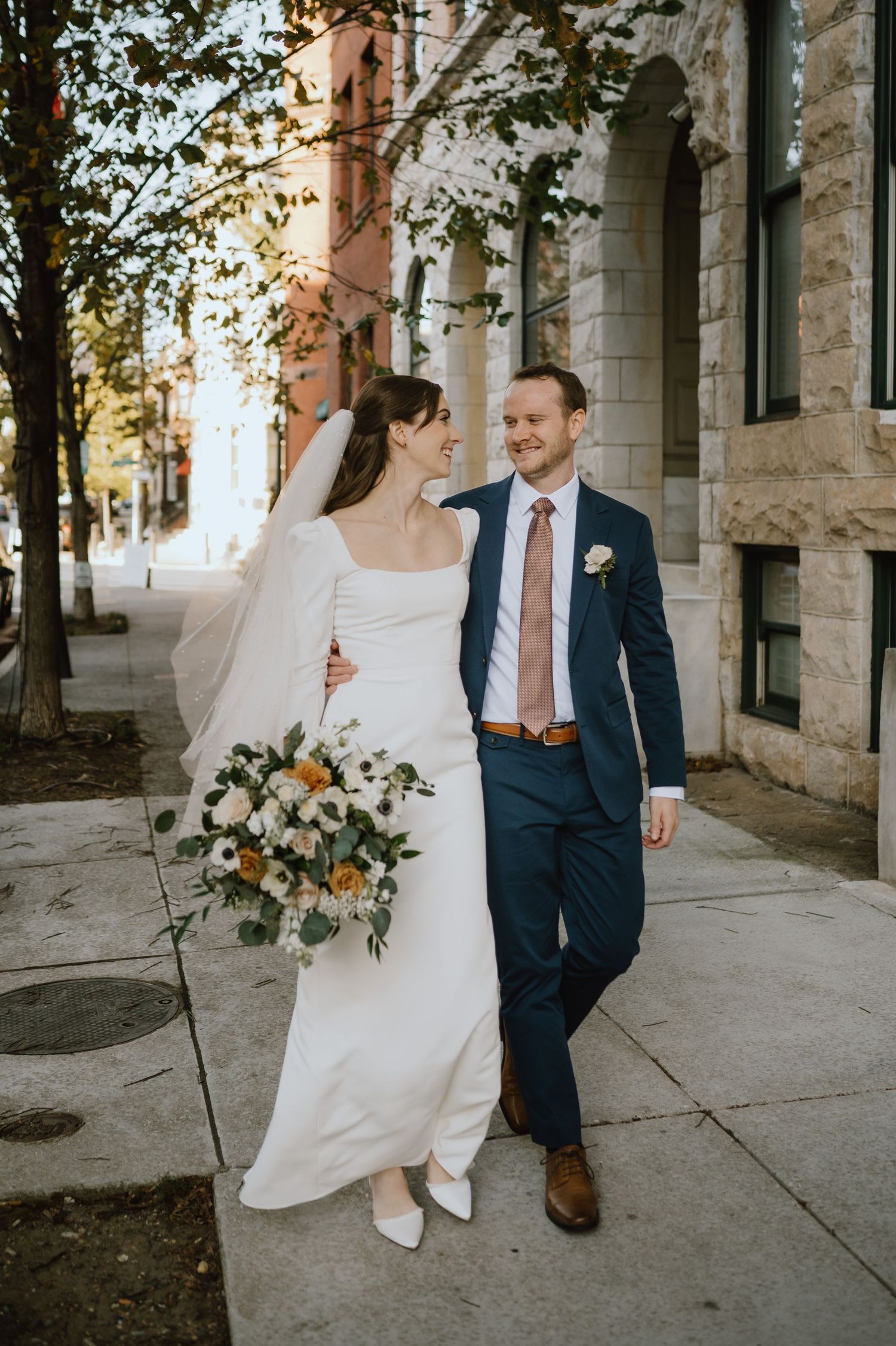 Couple walking on sidewalk with autumn trees and stone building in background.