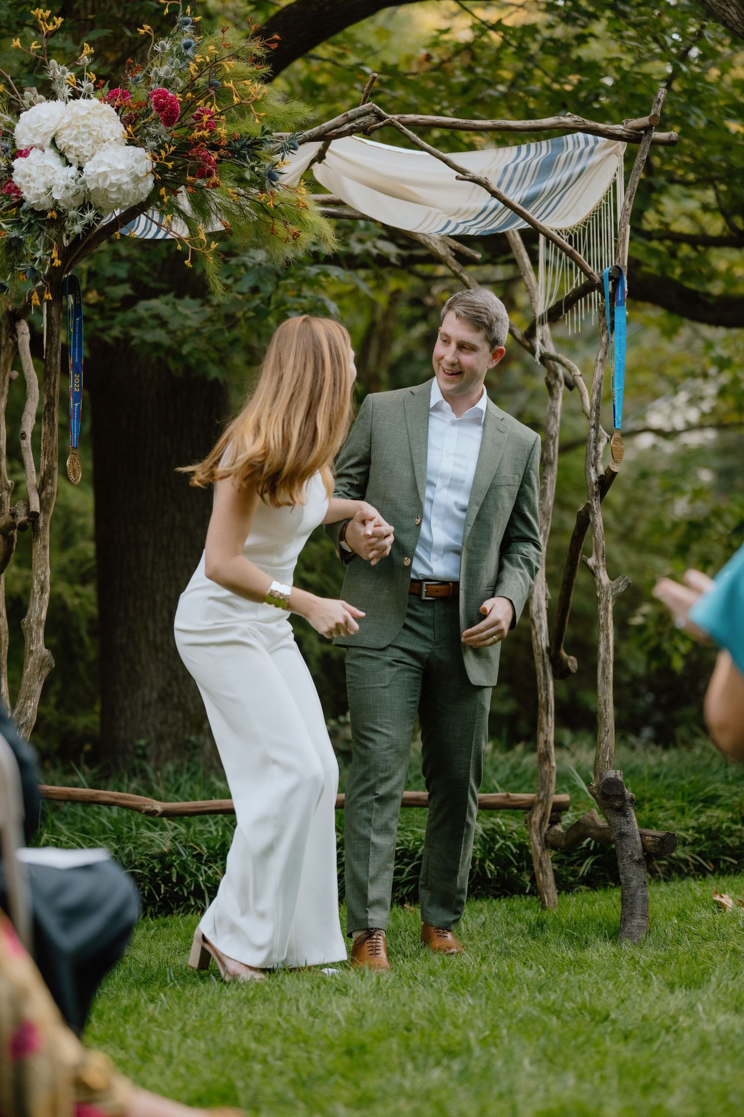 Elegant outdoor garden wedding ceremony with couple standing under rustic wooden arch decorated with white flowers.