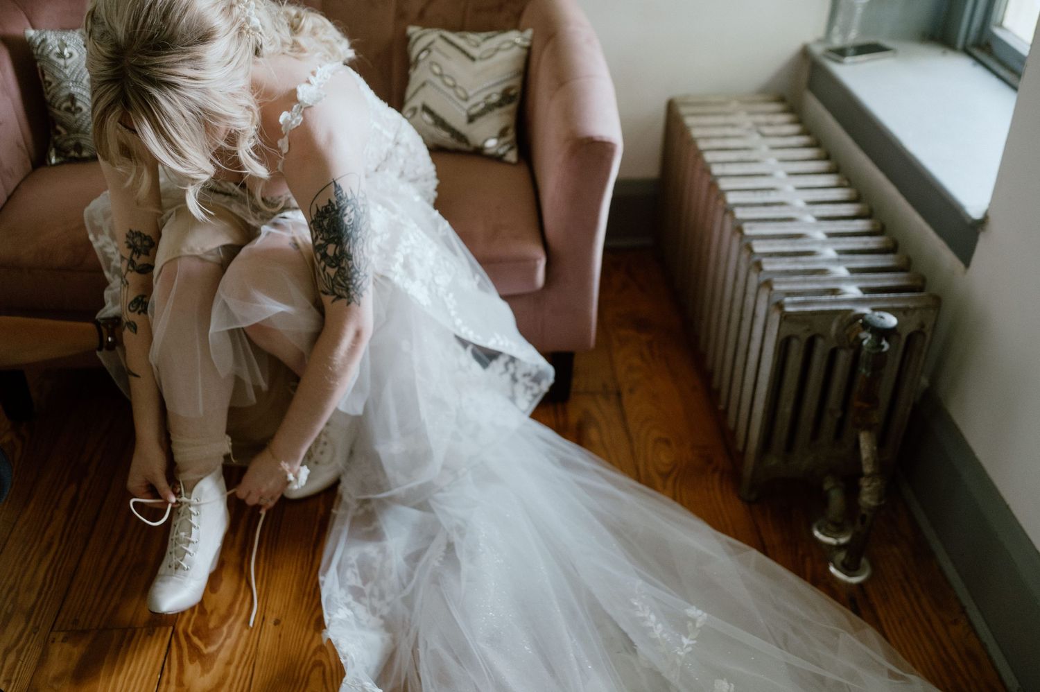 A person in a white wedding dress and sneakers rests on a brown couch near a window.