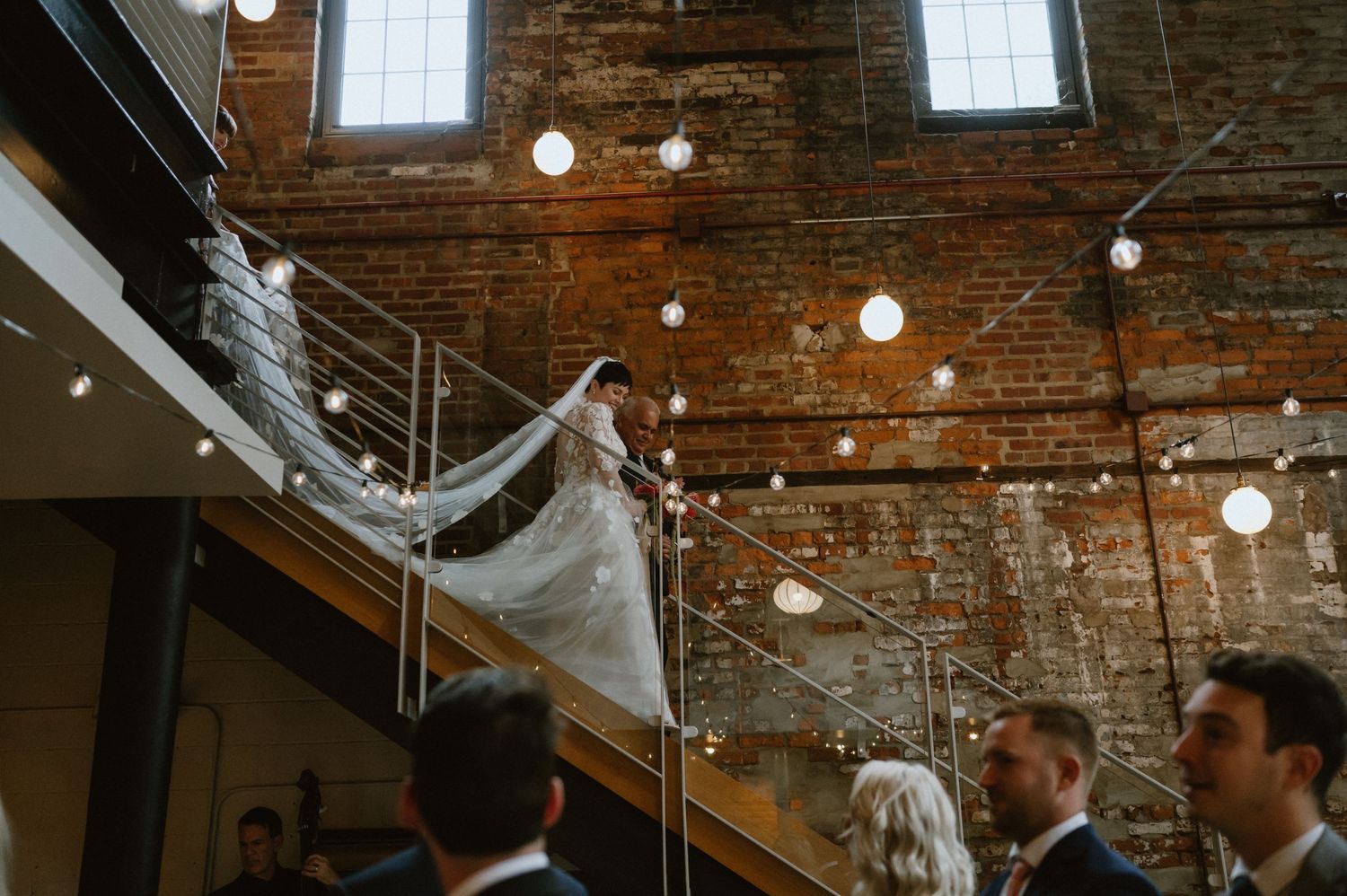Wedding guests gather beneath string lights and exposed brick walls near a modern staircase.