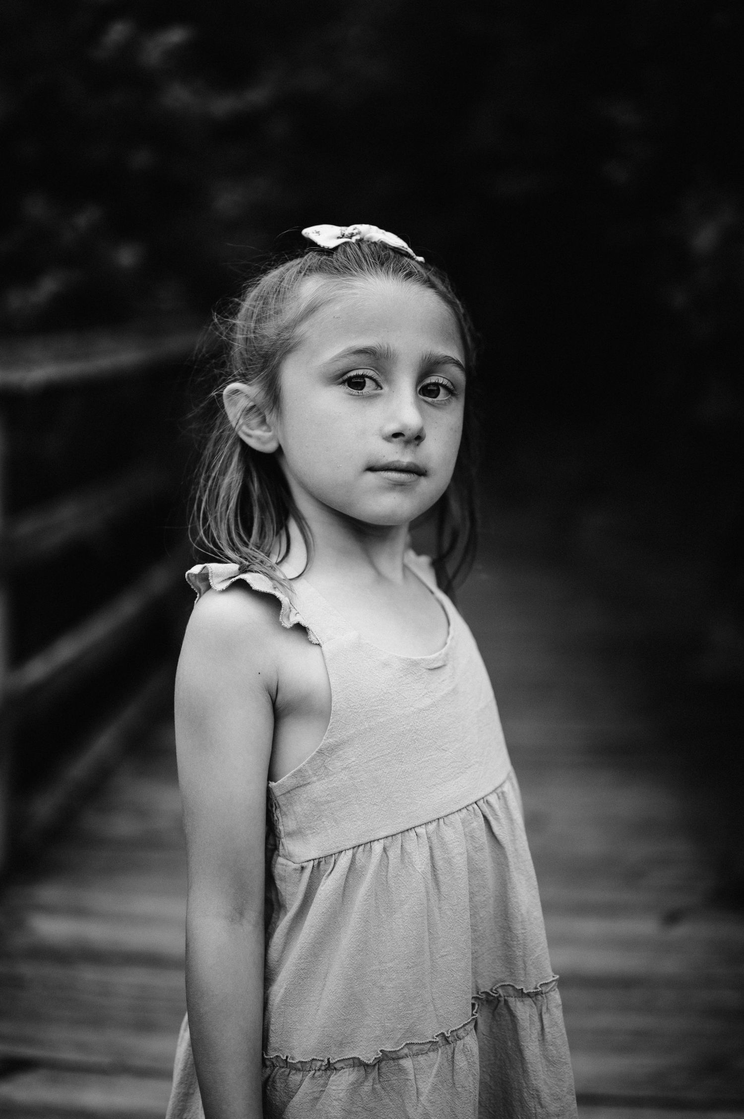 Portrait of child in light dress with hair bow standing on wooden boardwalk.