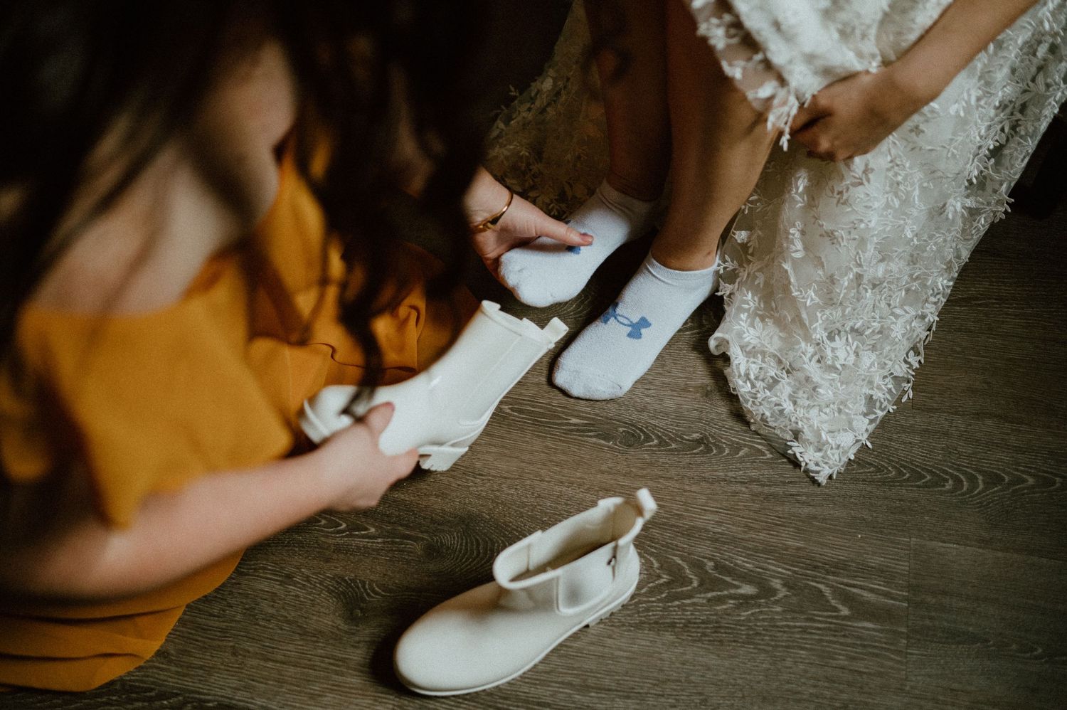Close-up of wedding shoes being fitted on wooden floor with lace dress details visible.