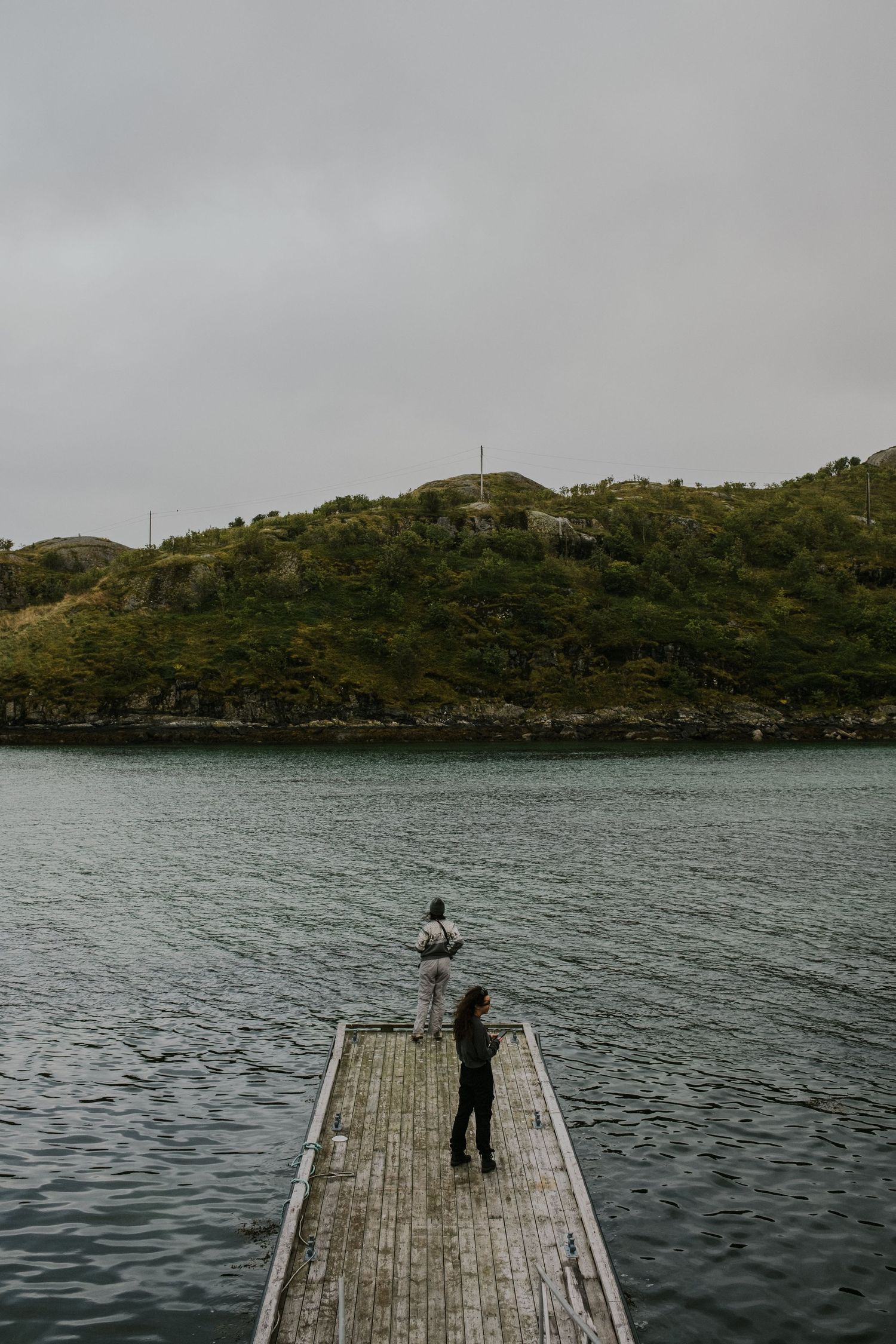 Figure standing at the end of a wooden pier overlooking tranquil waters and green hillside.