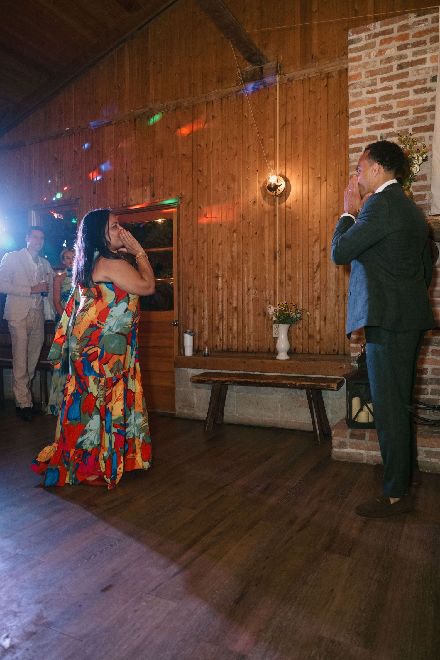 Wedding guests in colorful attire singing karaoke in a rustic wooden venue.