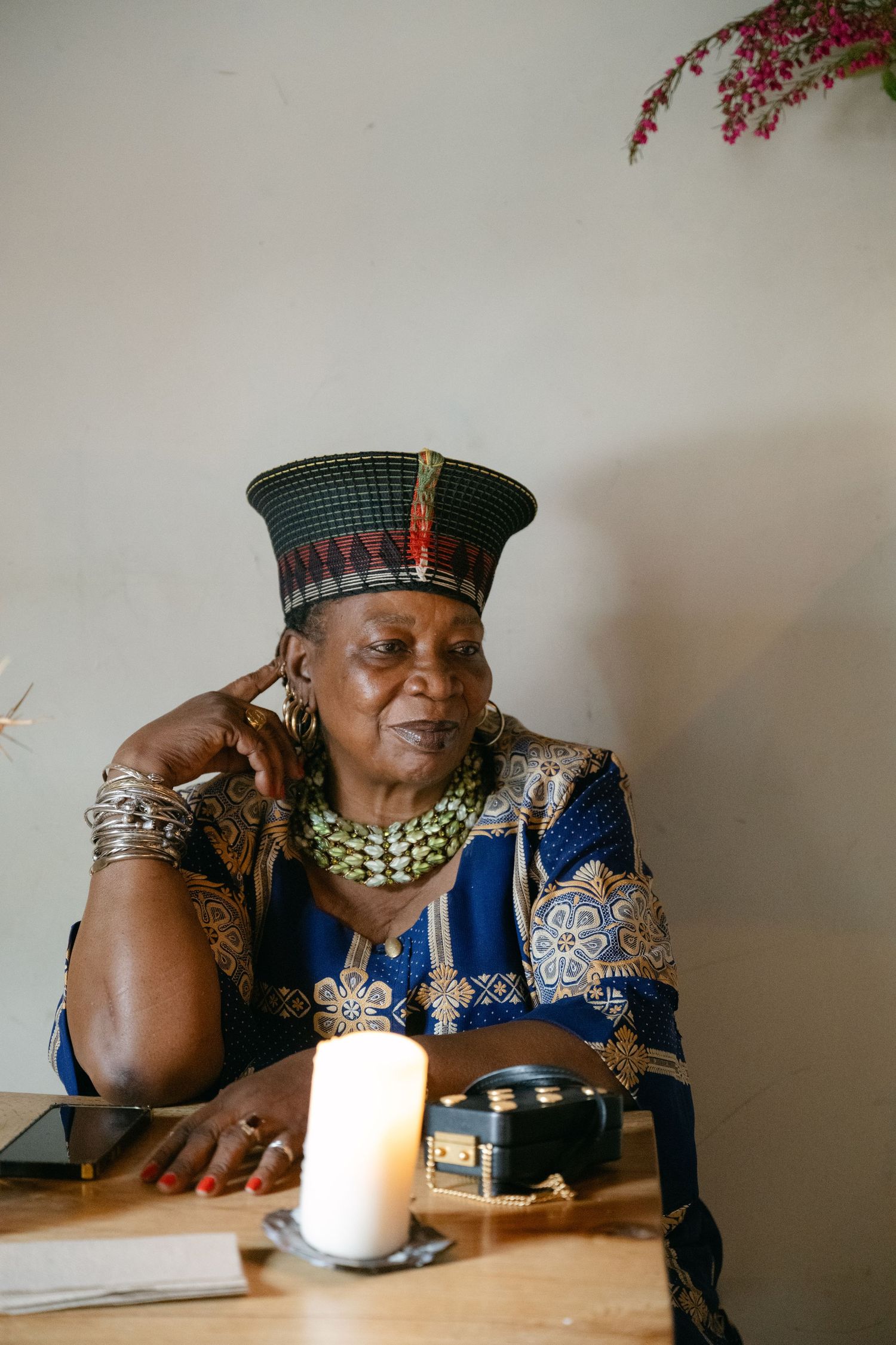 A person in traditional African attire and jewelry sits at a table with a candle while deep in thought.