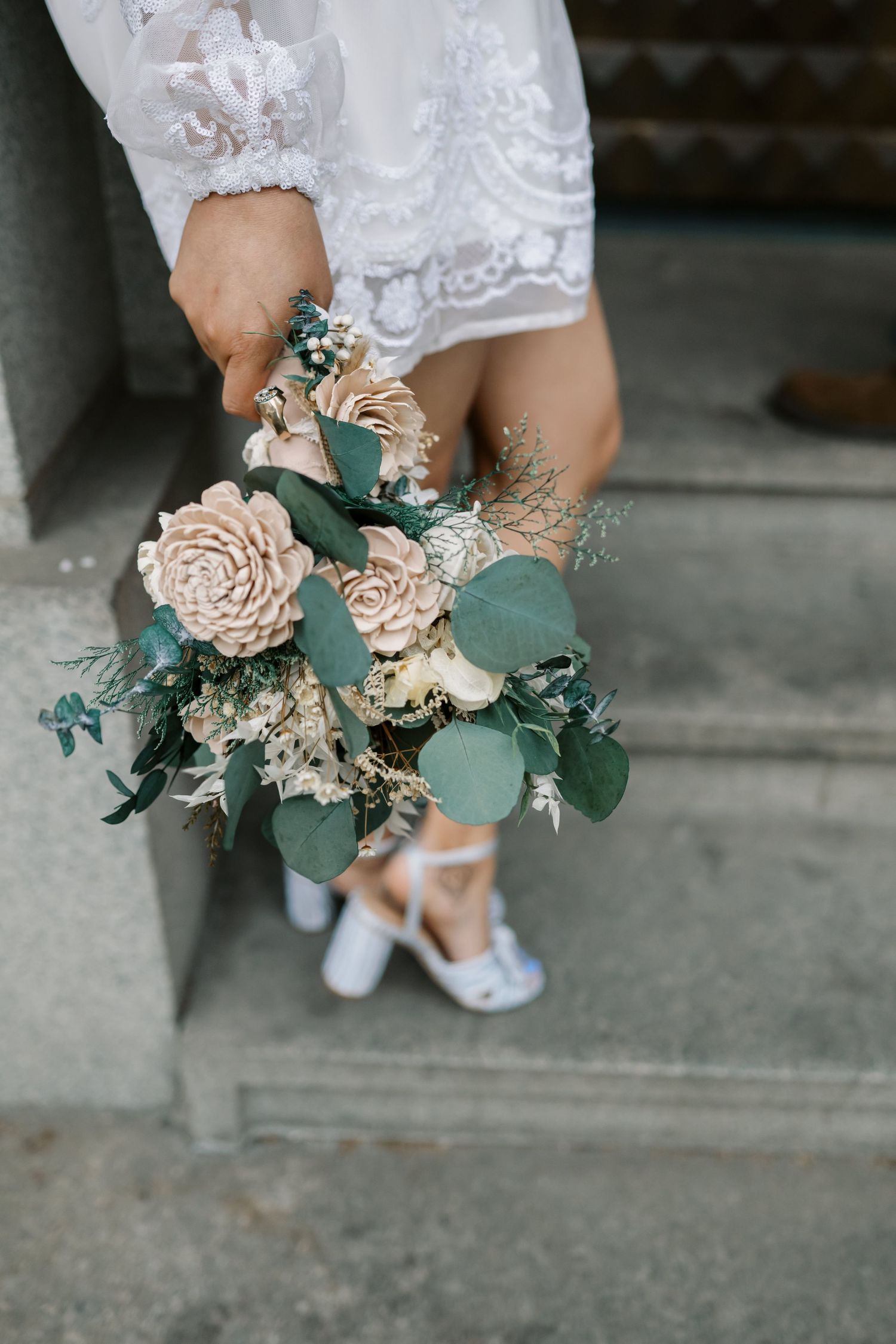 A delicate bridal bouquet of blush sola wood flowers and eucalyptus leaves held by someone in a lace dress and shoes.