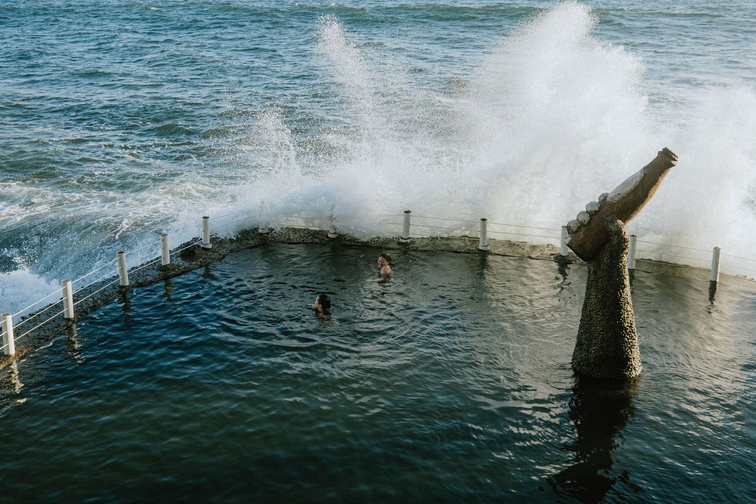 Waves crash over the edge of an ocean pool with stone barriers and sculptural posts while swimmers enjoy the water.