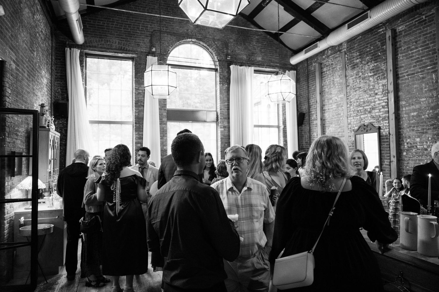 Black and white photo of reception guests mingling in historic venue with arched windows.