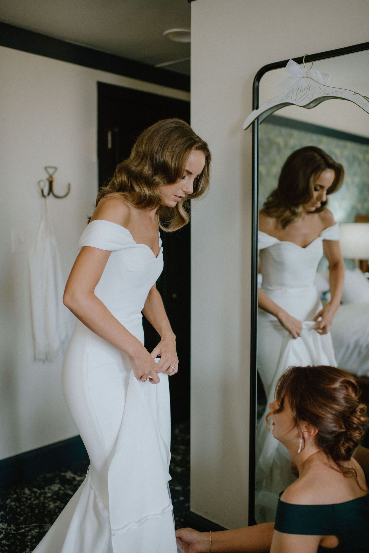 A bride tries on her elegant off-shoulder wedding gown in front of a mirror.