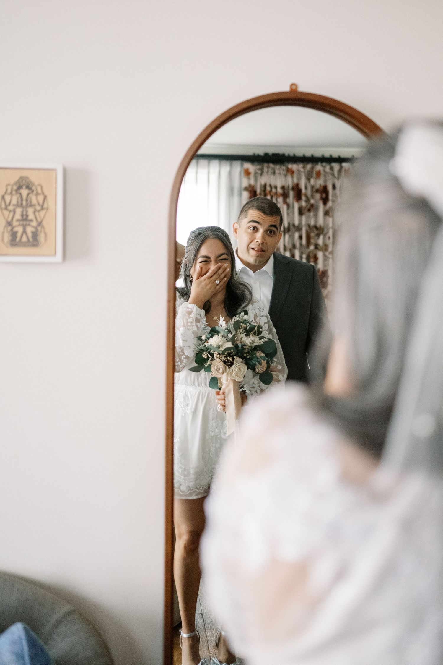 A couple in wedding attire sharing a joyful moment while looking at their reflection in a round wooden-framed mirror.