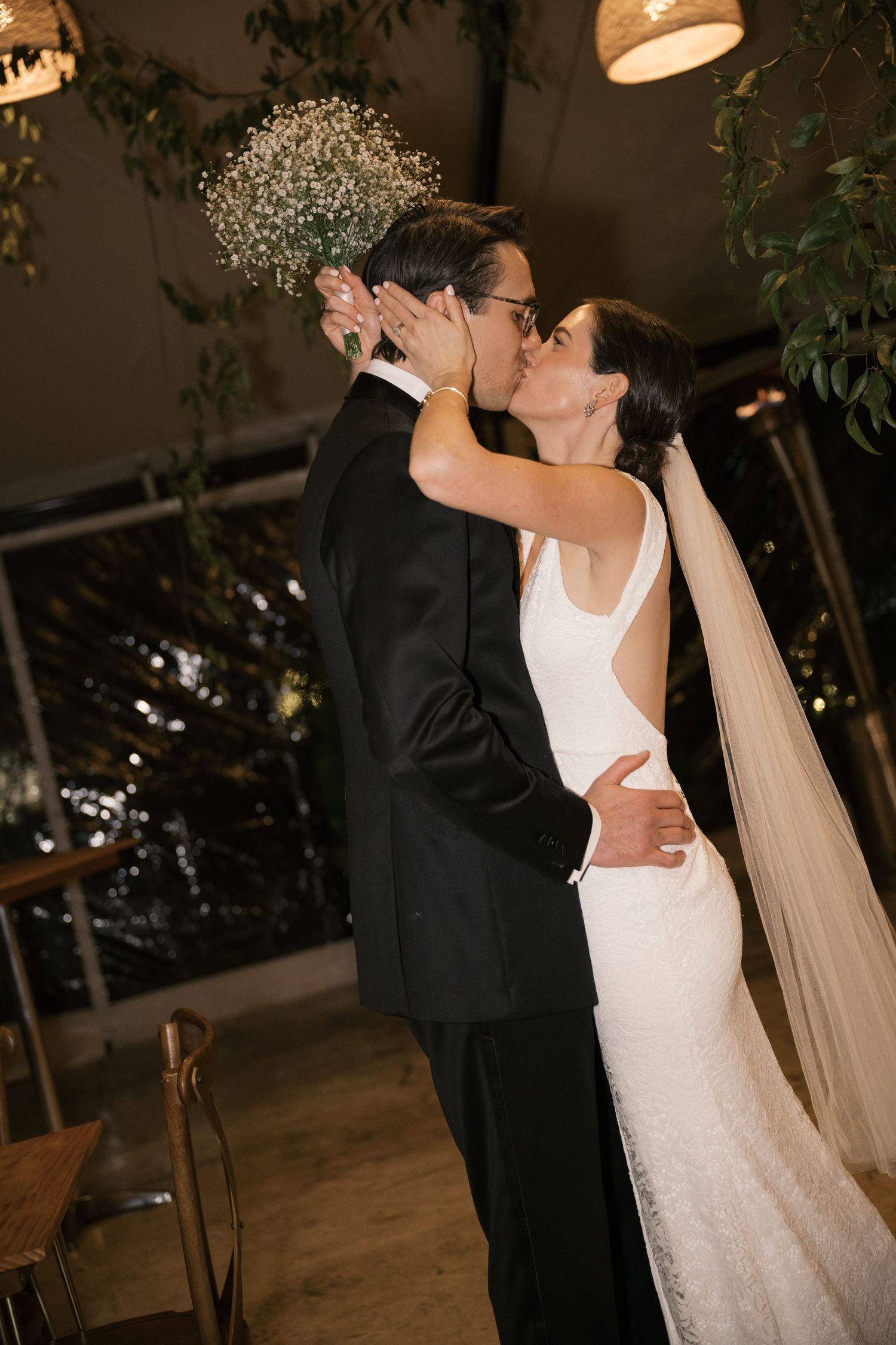 A romantic wedding moment showing a bride in white and groom in black sharing a kiss in an indoor venue with greenery.