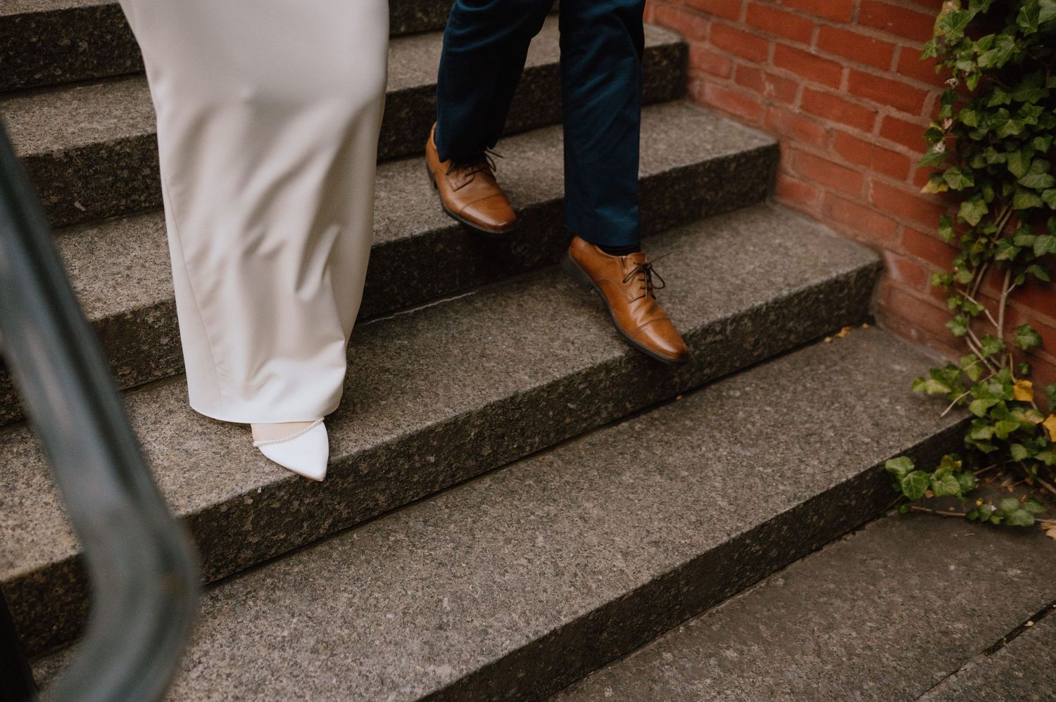 Wedding couple stands on steps against an ivy-covered brick wall.