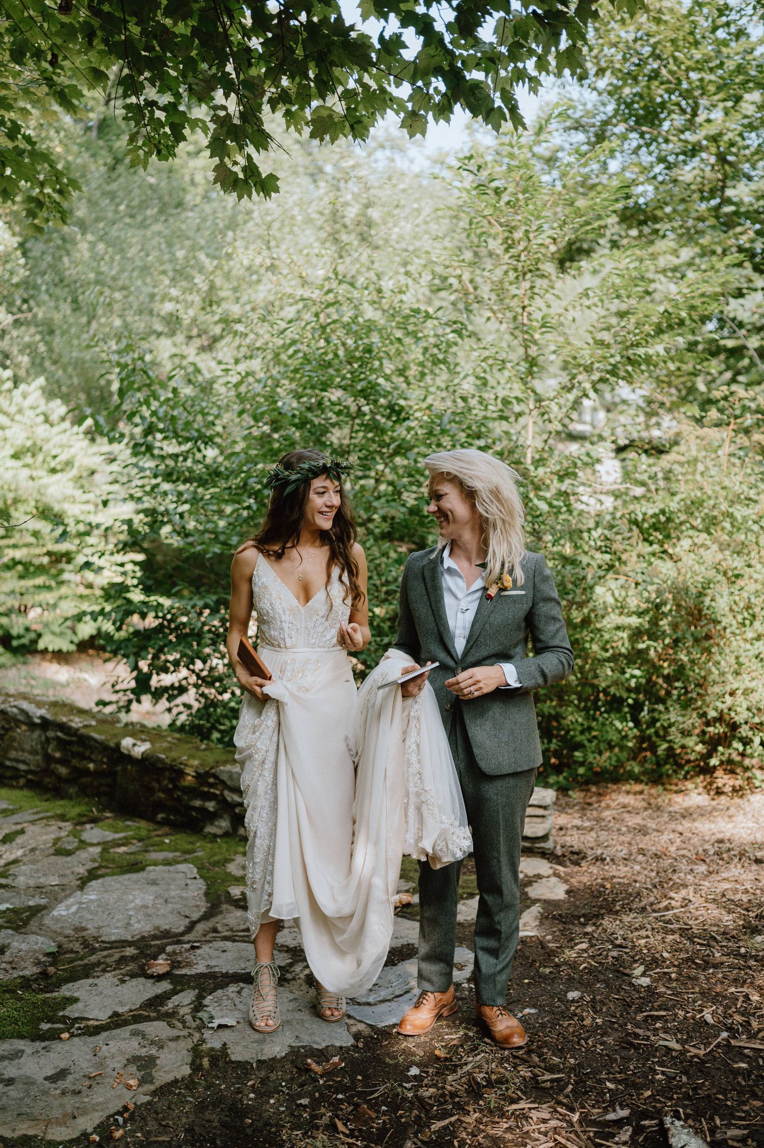 Two people in wedding attire share a joyful moment on a stone path surrounded by lush greenery.