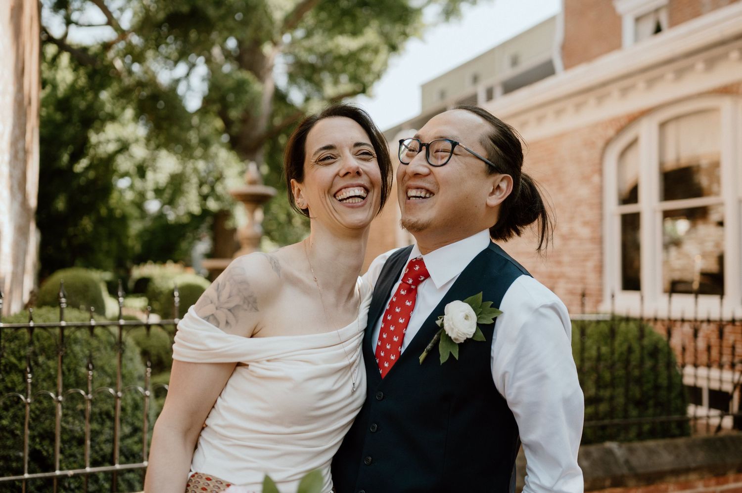 A joyful wedding couple laughs together outside a brick building on their special day.