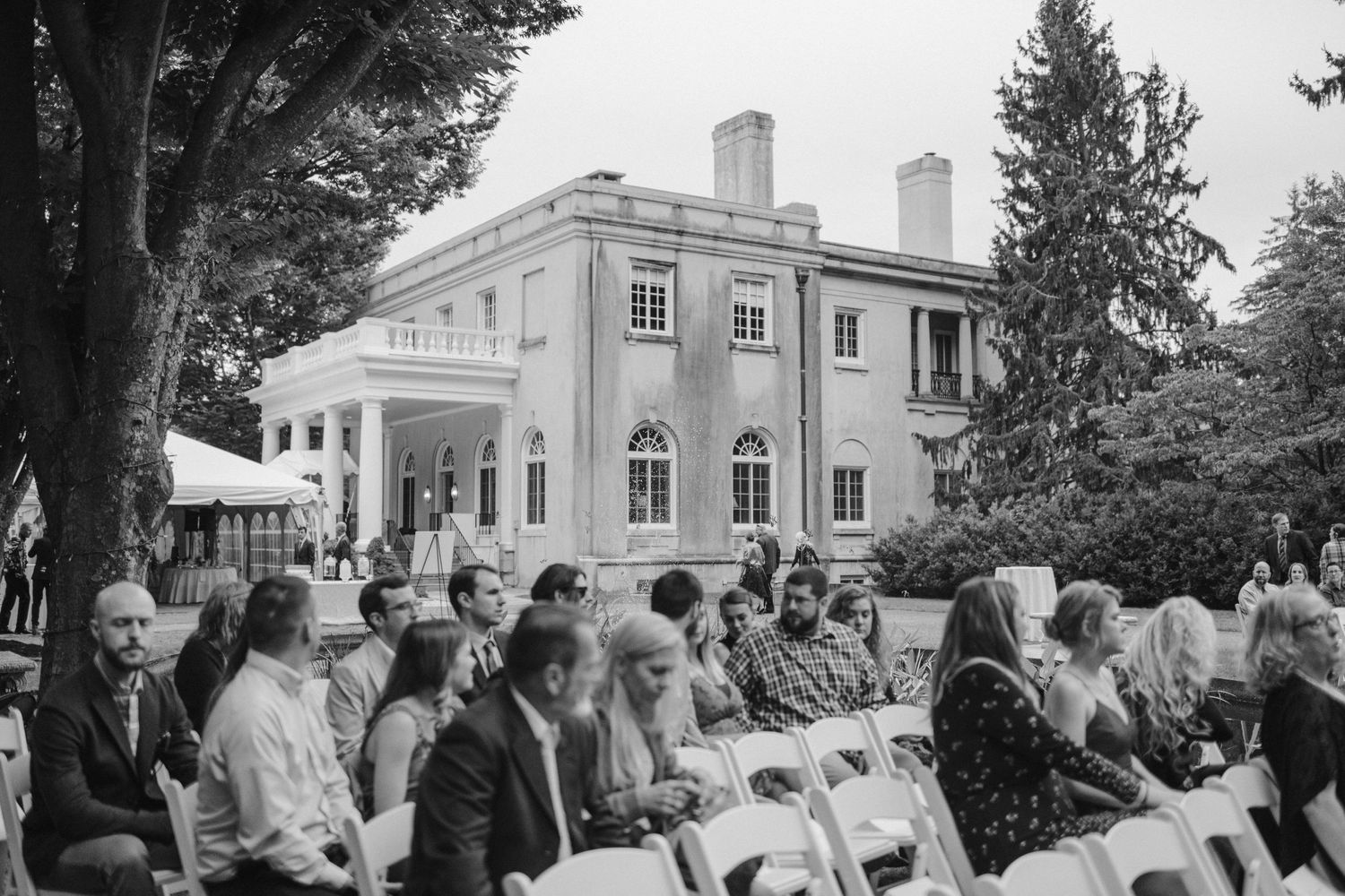 Large white mansion hosting an outdoor wedding ceremony with guests seated on white chairs.