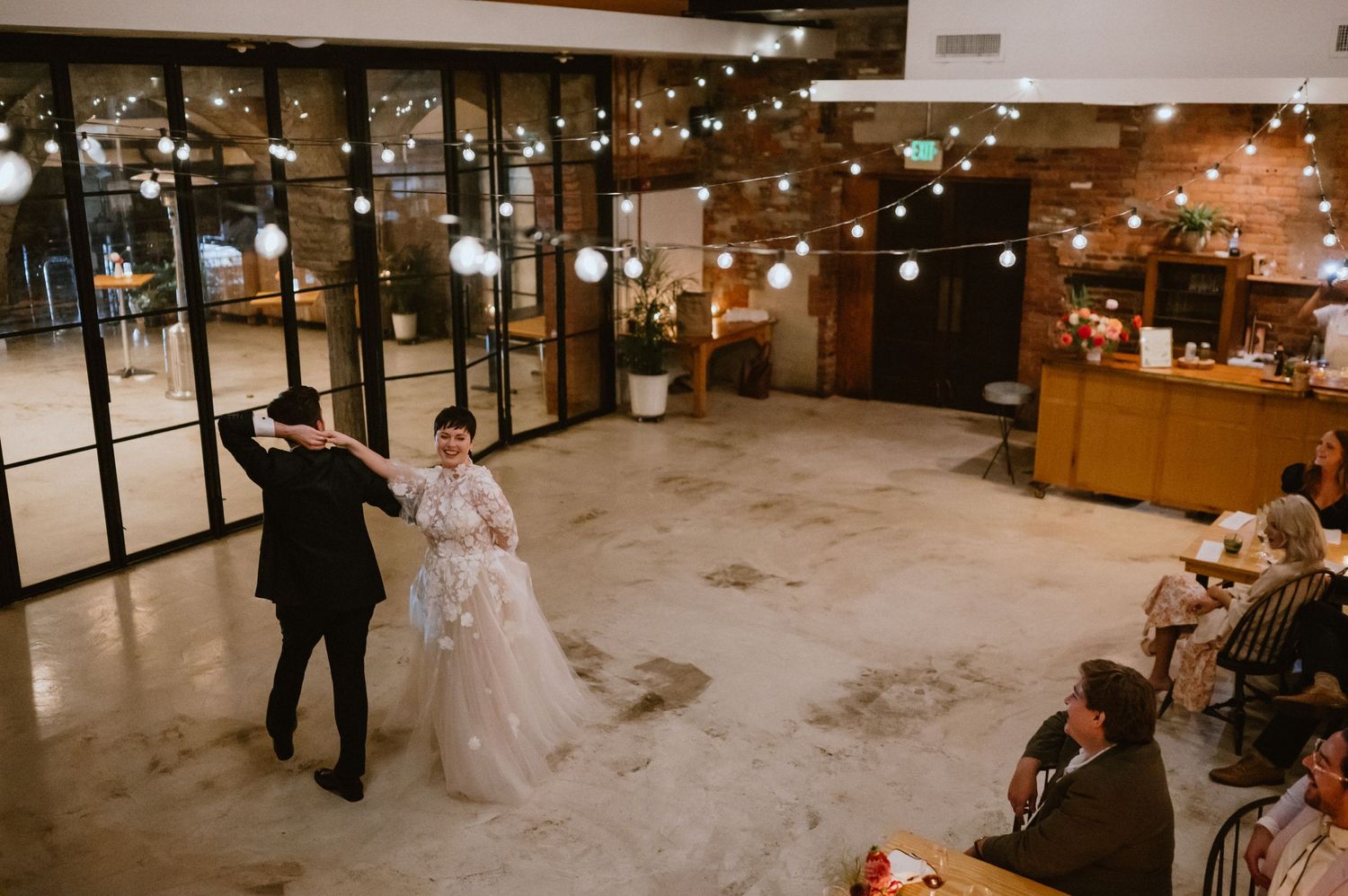 A couple dances their first dance in an industrial-style wedding reception venue with string lights overhead.