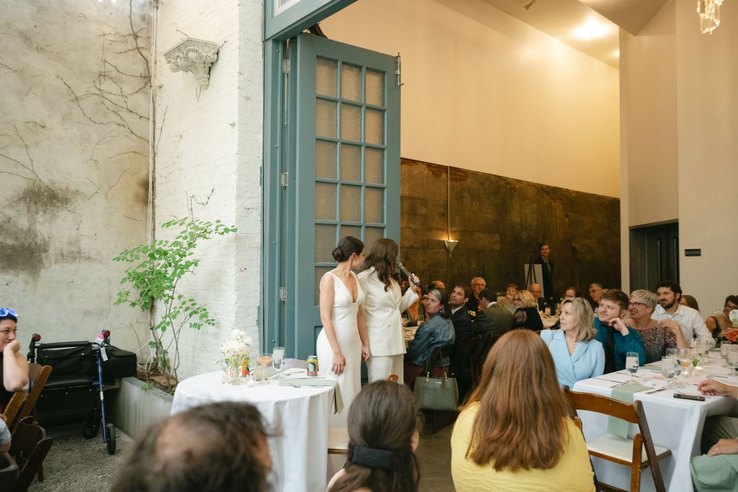 A couple stands at the front of a room with white tables while guests look on during an intimate indoor celebration.