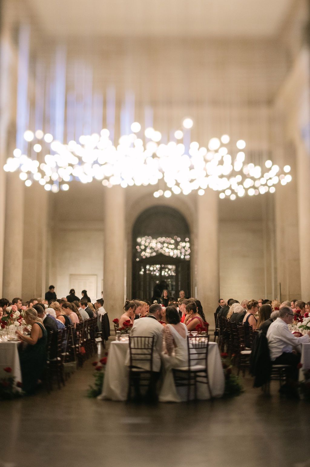 Elegant wedding reception venue with crystal chandeliers and long tables filled with guests in a white columned ballroom.